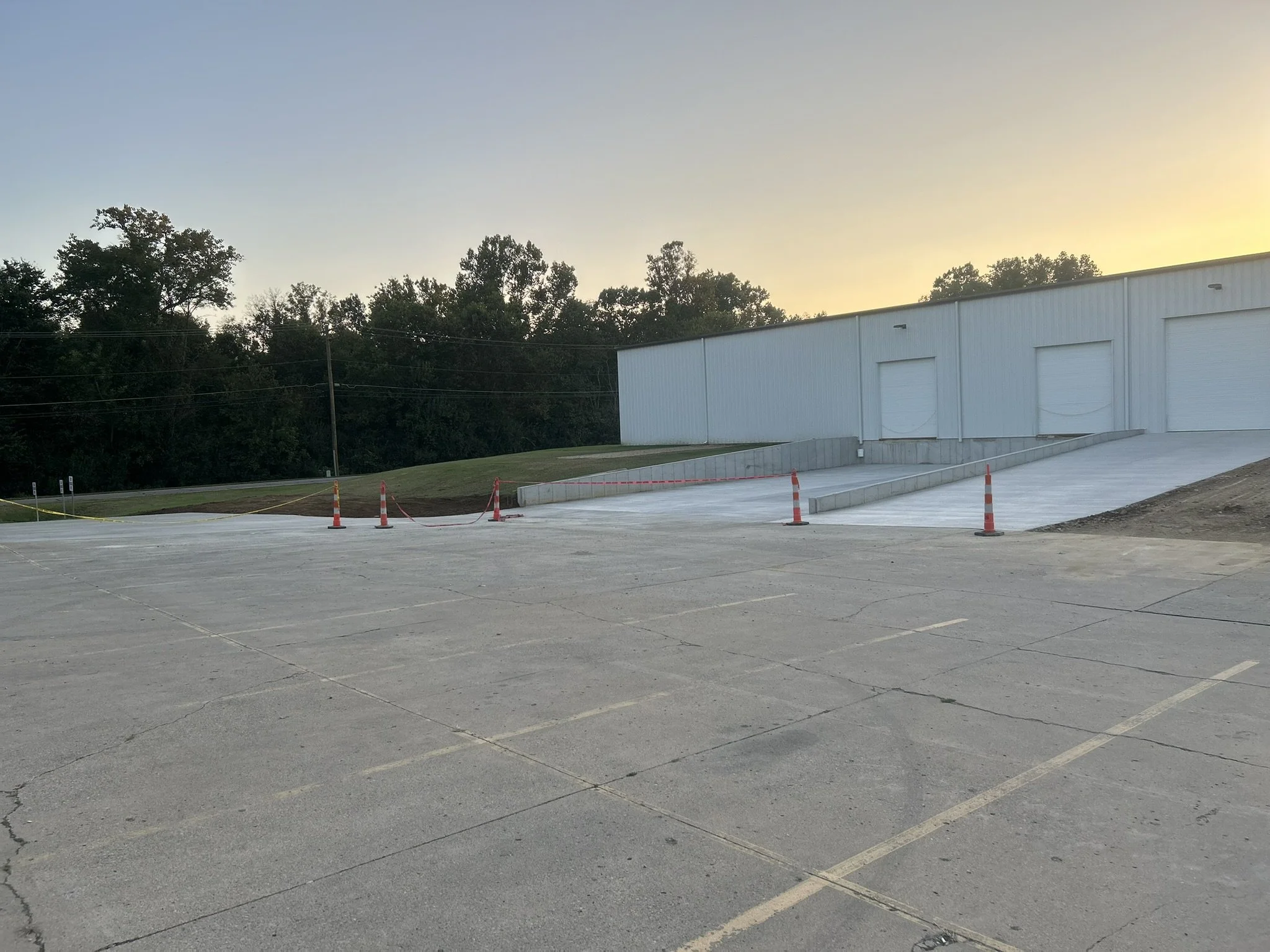 Empty parking lot with orange safety cones and a white industrial building at sunset, trees in the background.