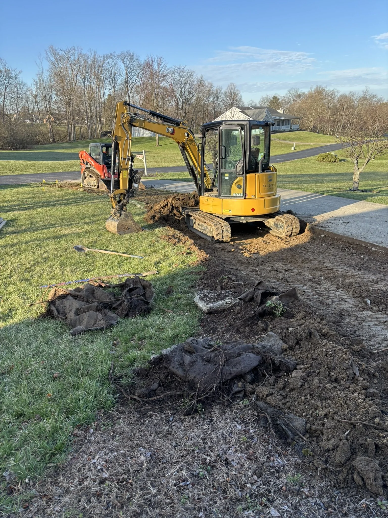 Excavator digging a trench in a grassy yard with trees and a house in the background.