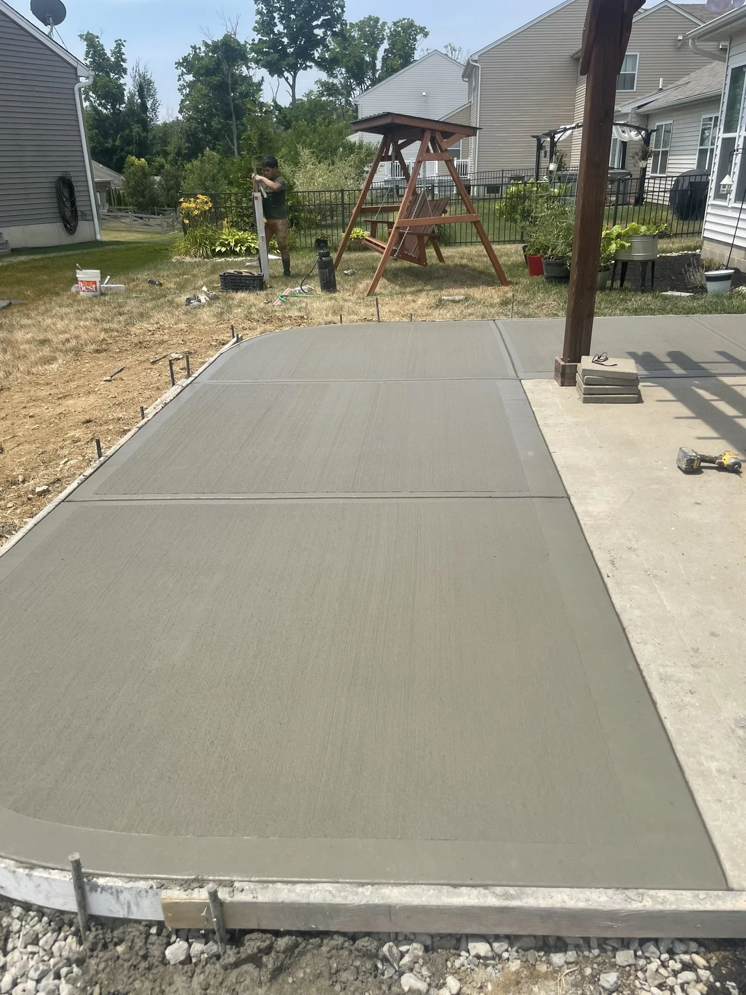 A newly poured concrete patio in a backyard with a wooden swing set, a person working with a level, and various gardening and construction tools and supplies.