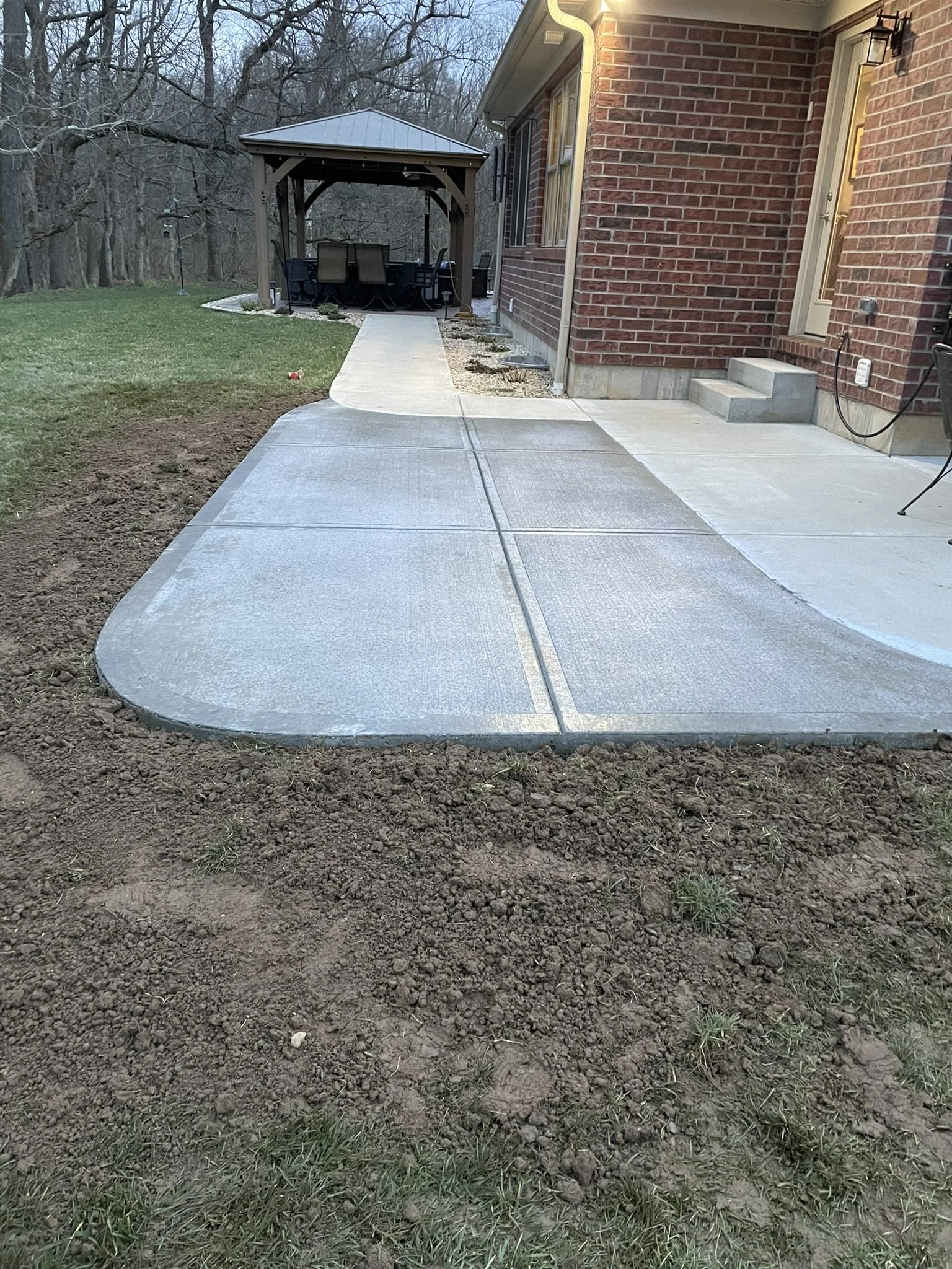 Concrete sidewalk leading to a backyard patio area with outdoor furniture and a gazebo, next to a brick house with steps and outdoor lighting.