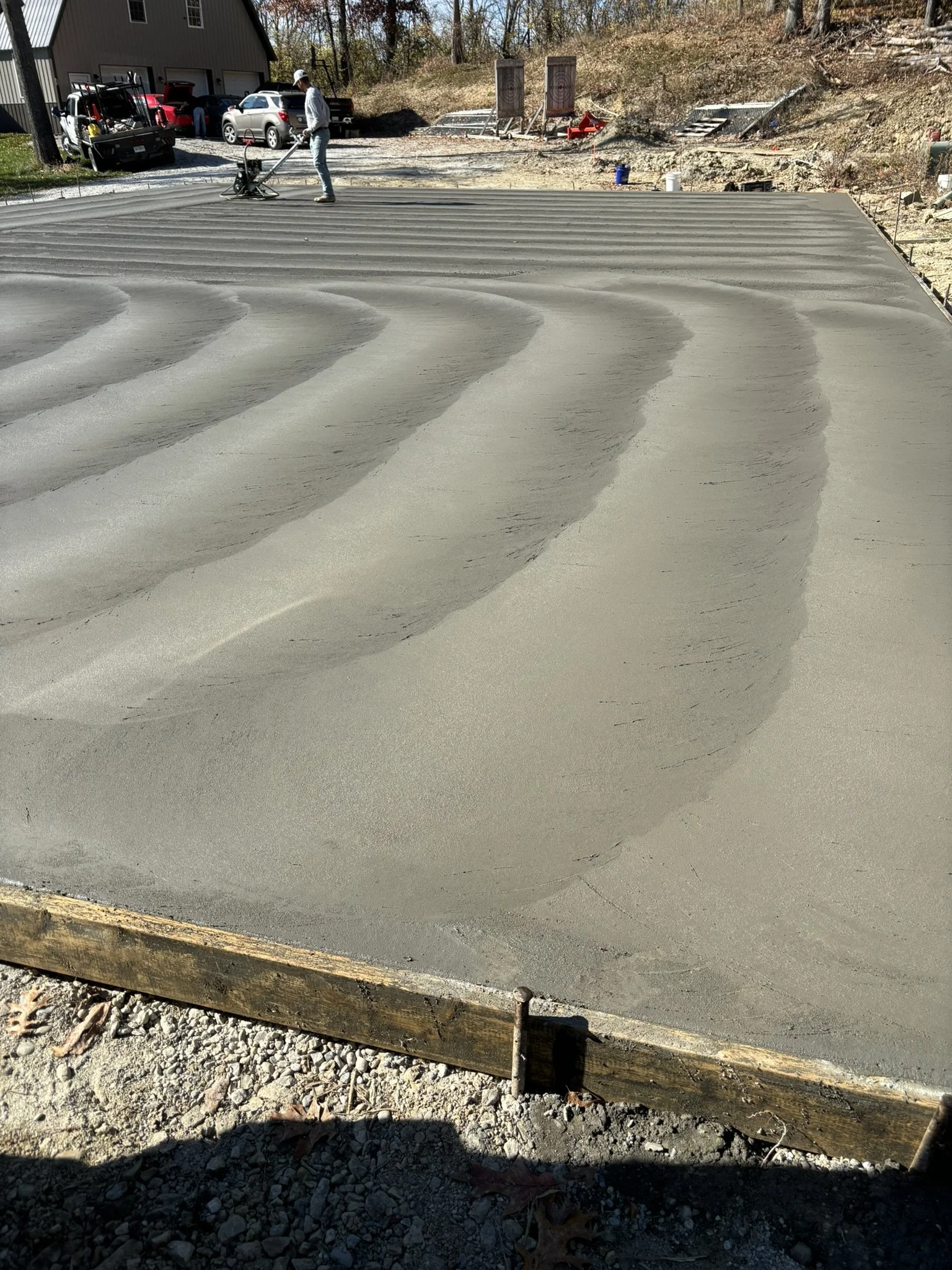 Worker smoothing freshly poured concrete on a construction site with a power trowel, with a building and vehicles in the background.
