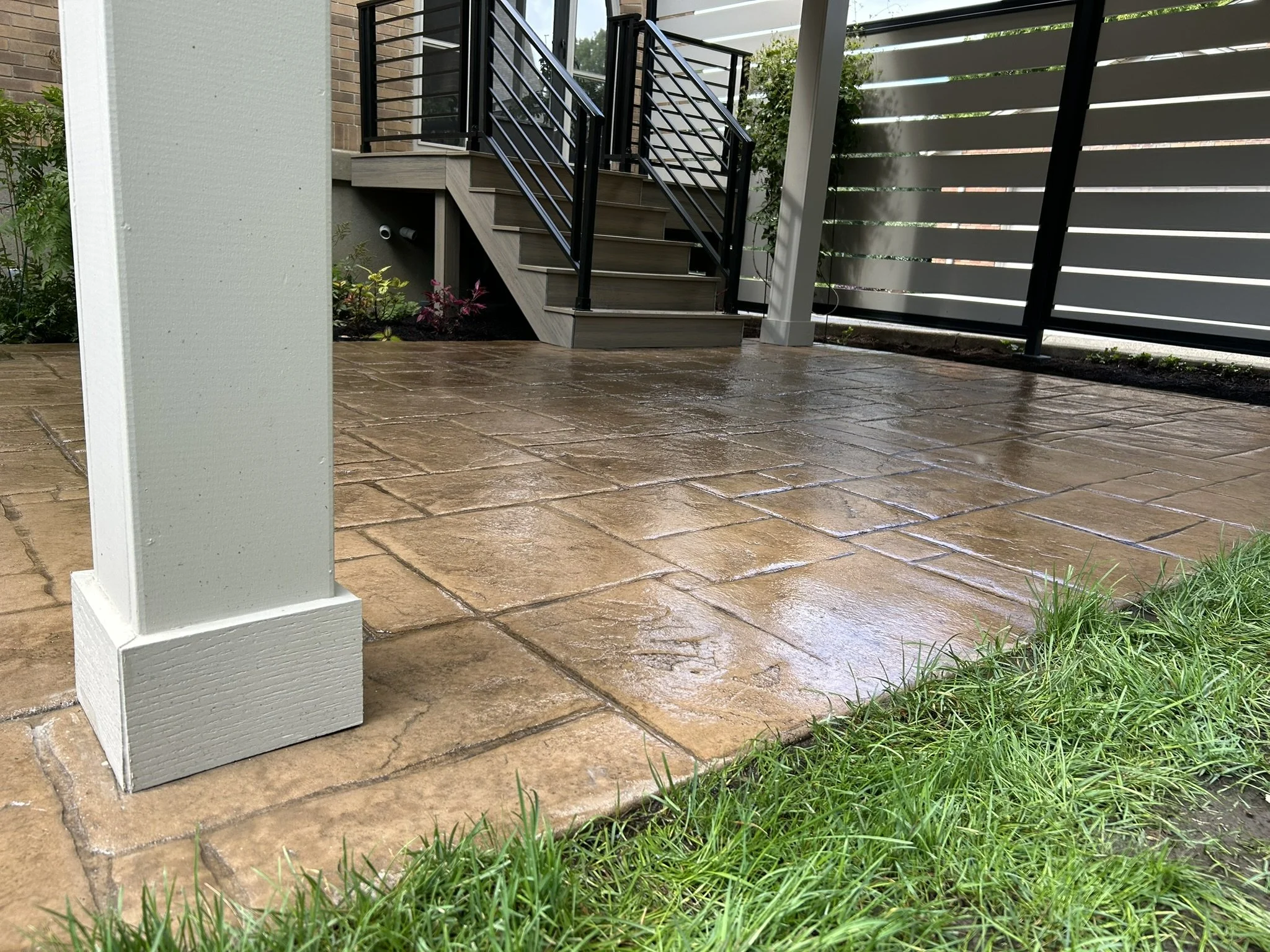 A backyard patio with wet brown stone tiles, a white painted column, a black metal staircase with wood steps, and a white fence with black posts. Green grass is visible at the edge of the patio.
