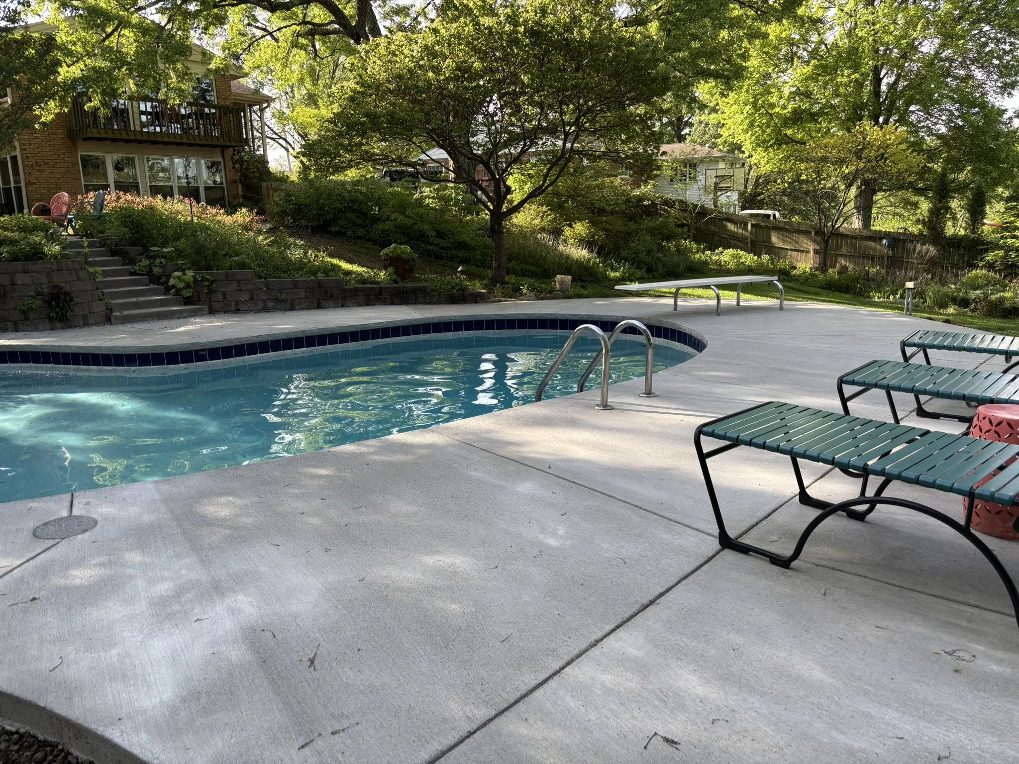 Backyard with a swimming pool, lounge chairs, a patio area, trees, and residential houses in the background.