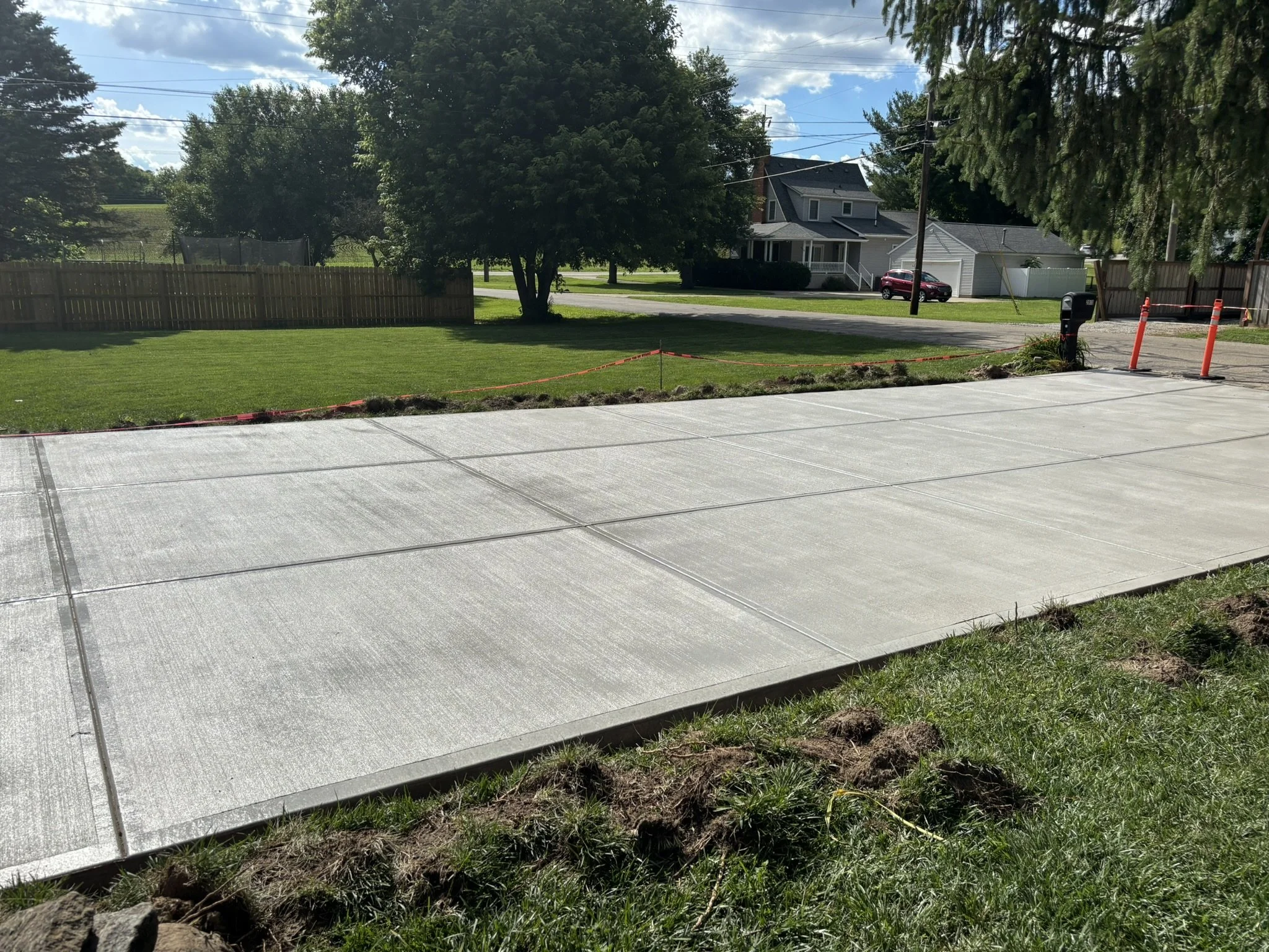 A newly poured concrete driveway with expansion joints, adjacent to a grassy lawn and flower bed, in front of a residential house with trees, power lines, and a parked car in the background.