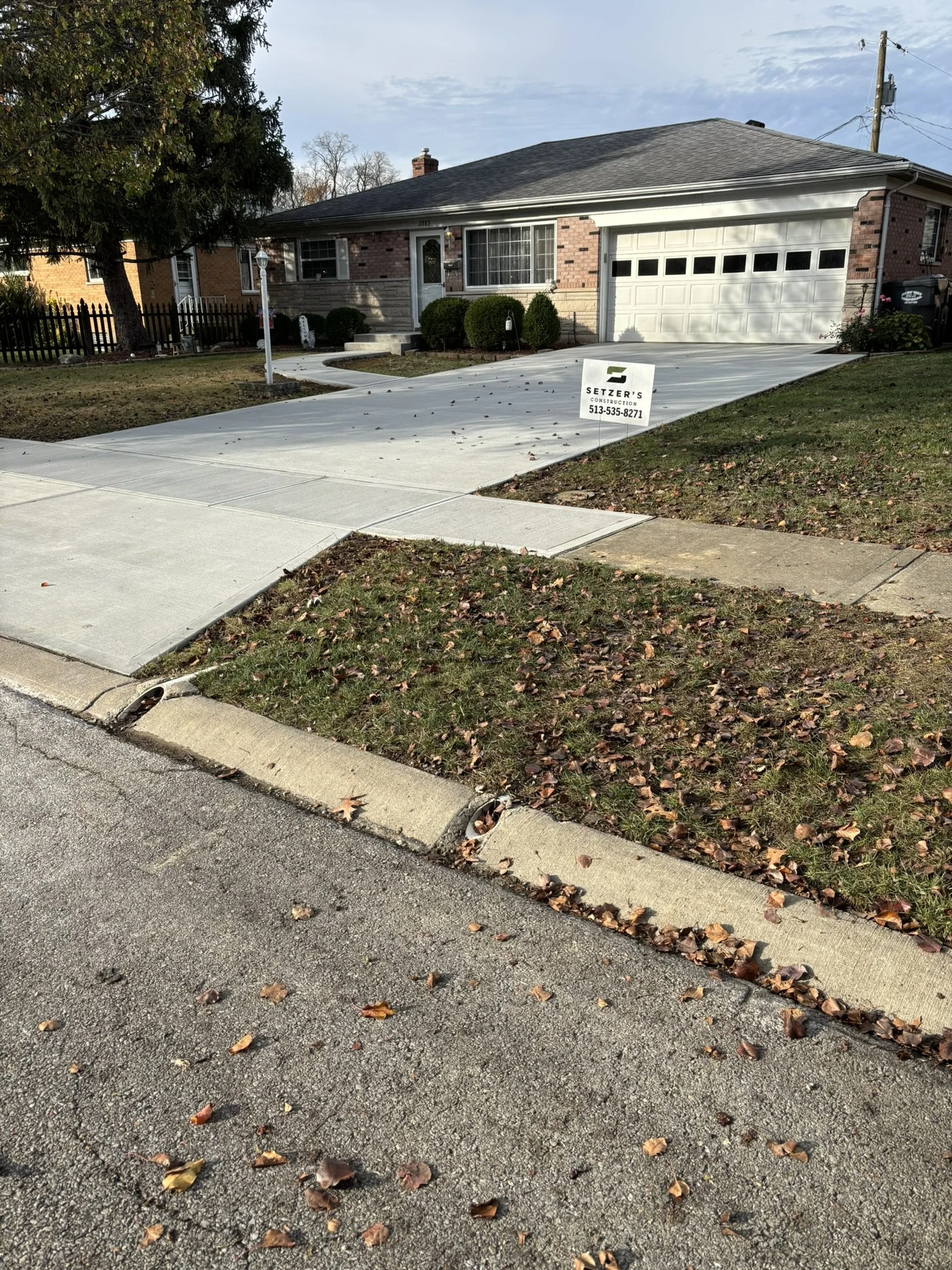 A house with a driveway, a lawn with fallen leaves, shrubs in front, and a sign for construction company Setzer's Construction. The house has a brick facade, a window with white curtains, and a two-car garage. The driveway is new, clean, and curved, 