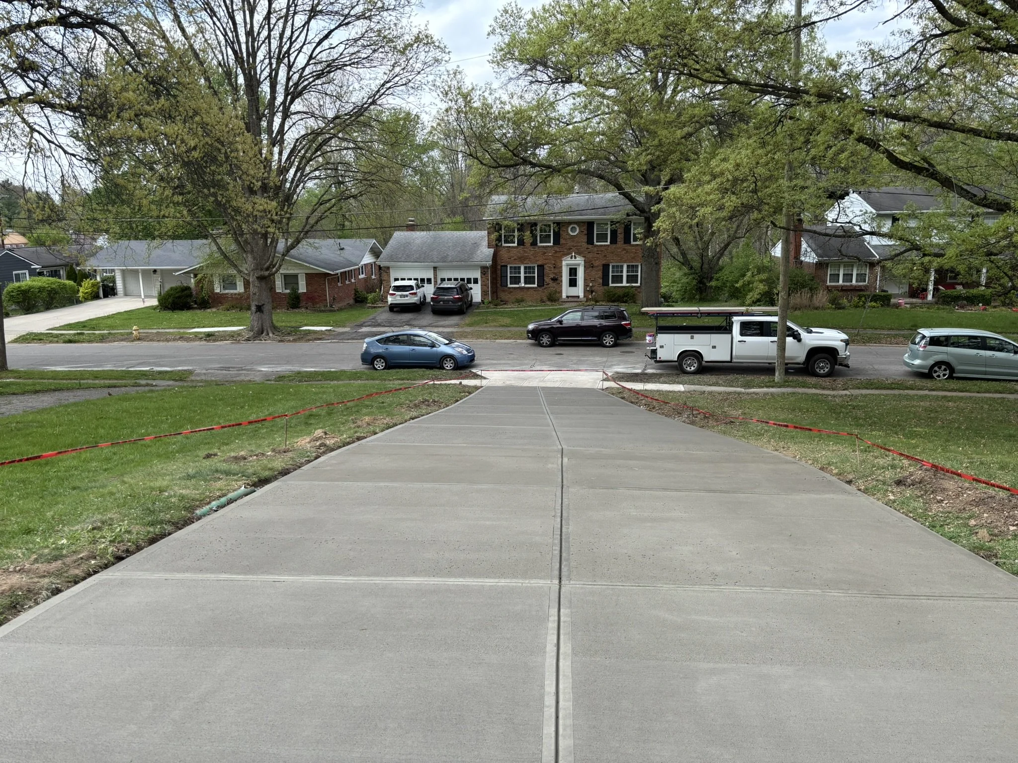 View of a suburban residential street with houses, large trees, cars parked along the curb, and a sidewalk leading down the stairs.