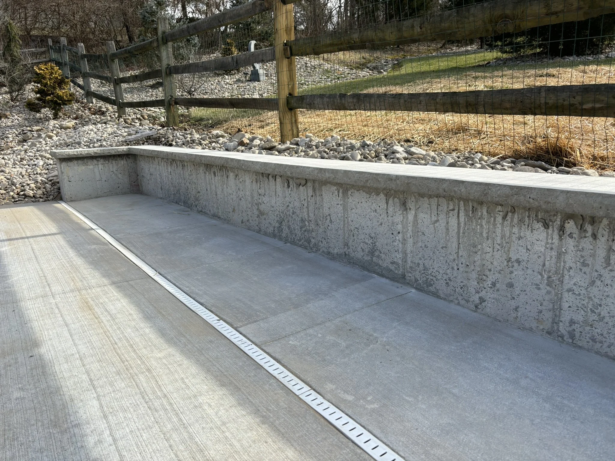 A concrete ledge beside a sidewalk, with a metal drain grate along its edge, a wooden and wire fence, and a sloped grassy area with rocks and some small bushes in the background.