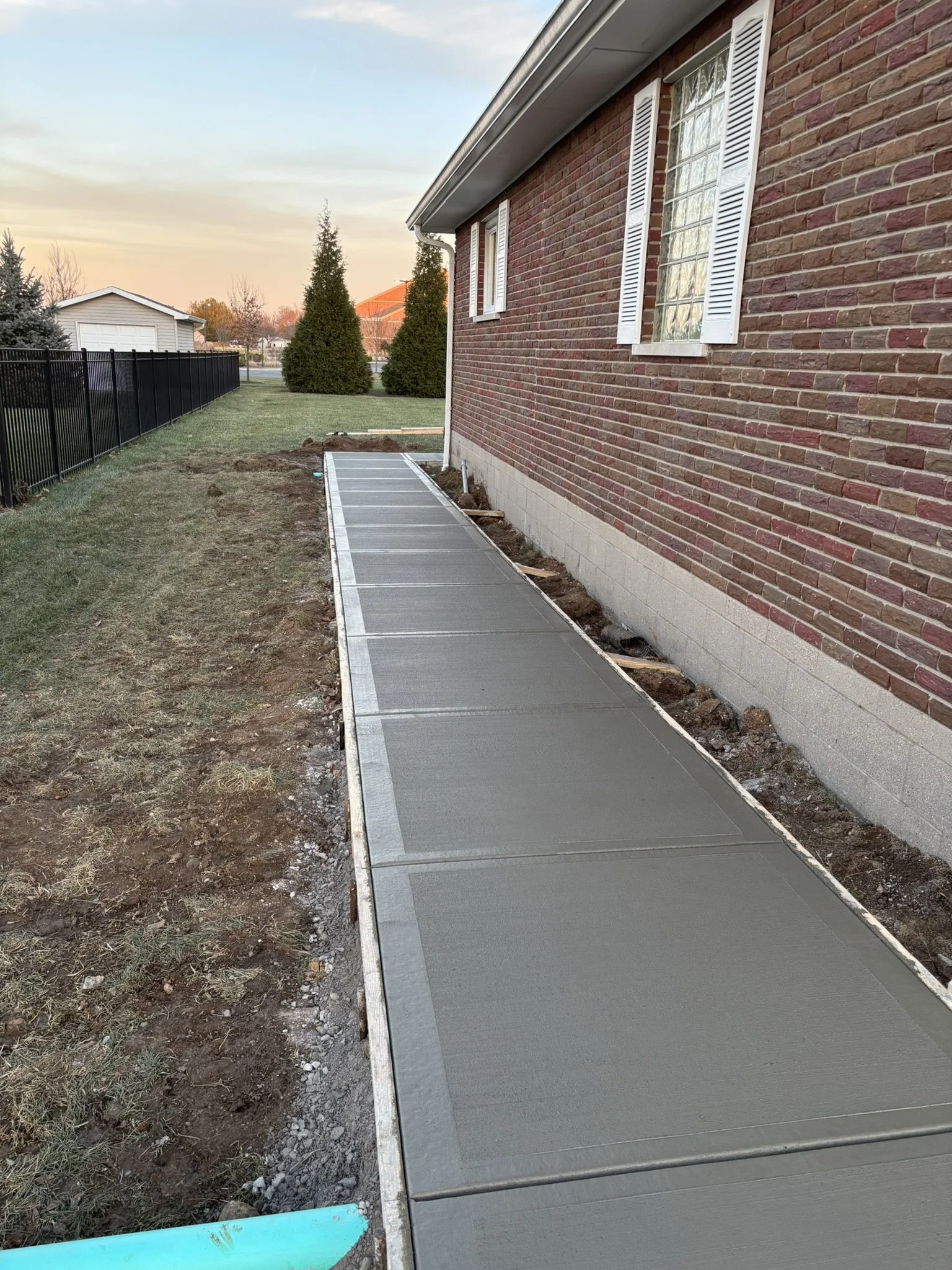 A newly paved concrete sidewalk runs alongside a brick house with white window shutters, with a grassy yard on one side and dirt on the other, during sunset.