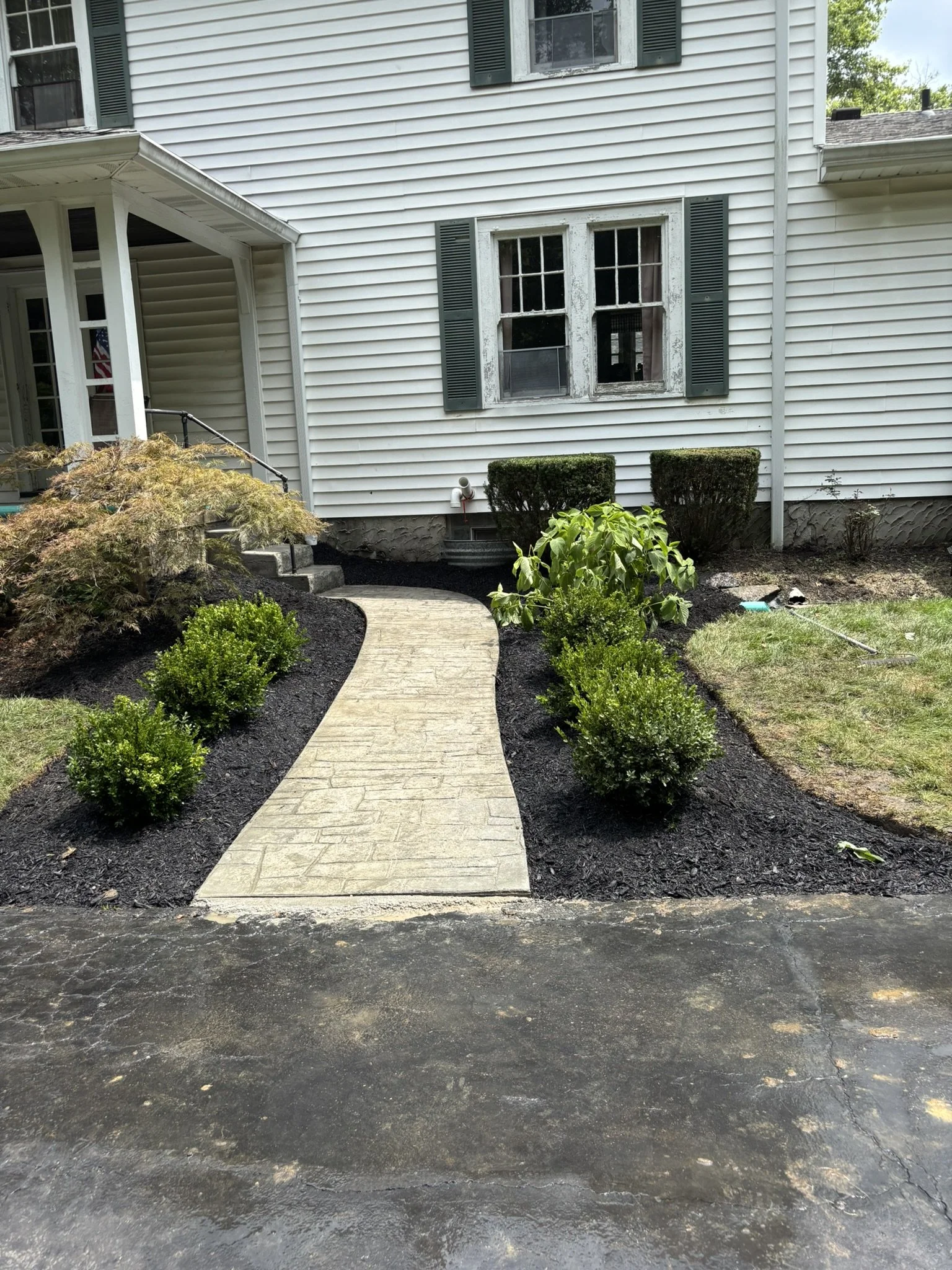 Side view of a house with white siding, a window with dark green shutters, a small front yard with a stone pathway, bushes, and freshly mulched garden beds, with a freshly paved driveway in the foreground.