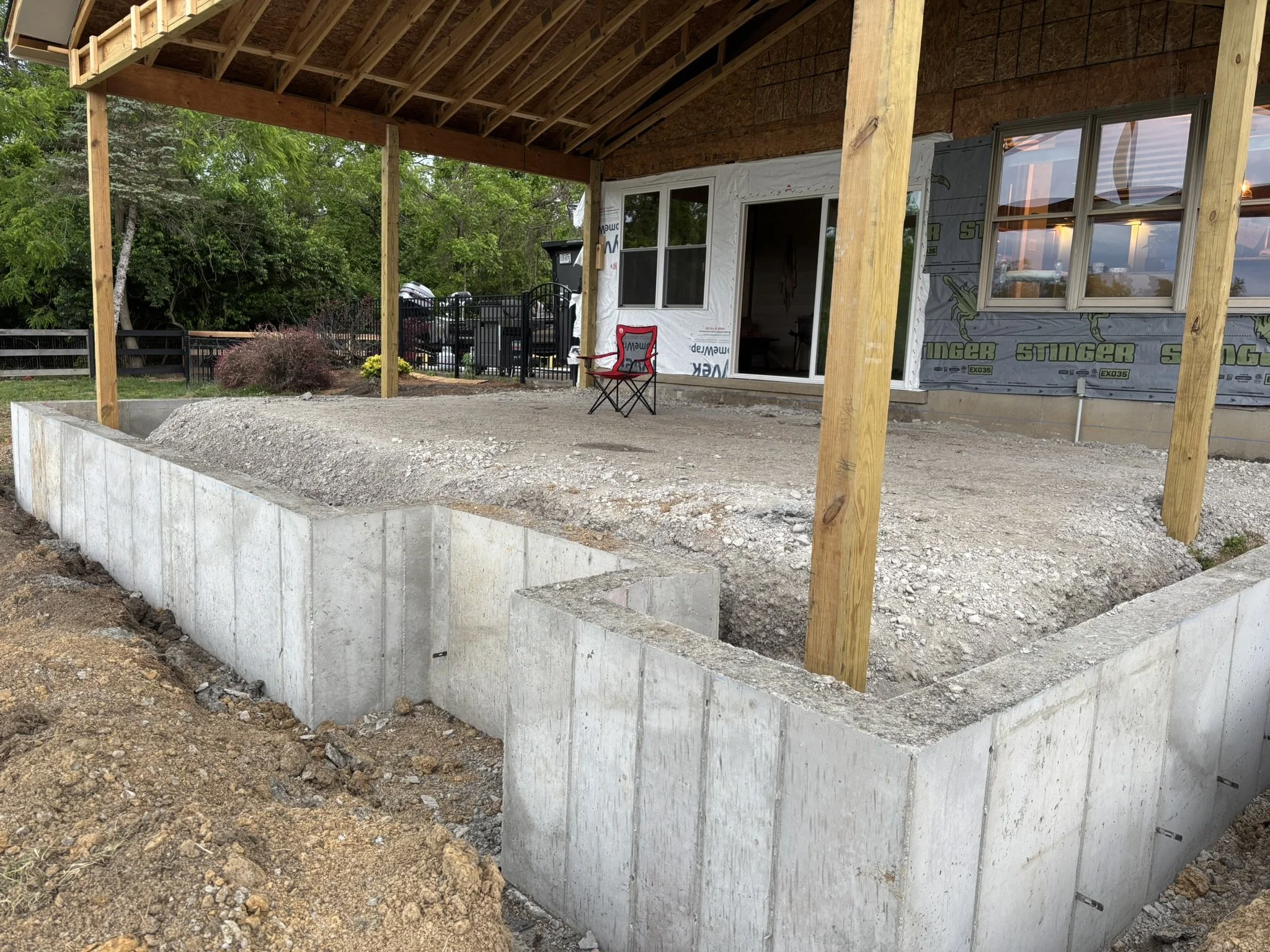Construction site with a concrete foundation, wooden support beams, and an unfinished house with a porch, surrounded by trees and a black fence.
