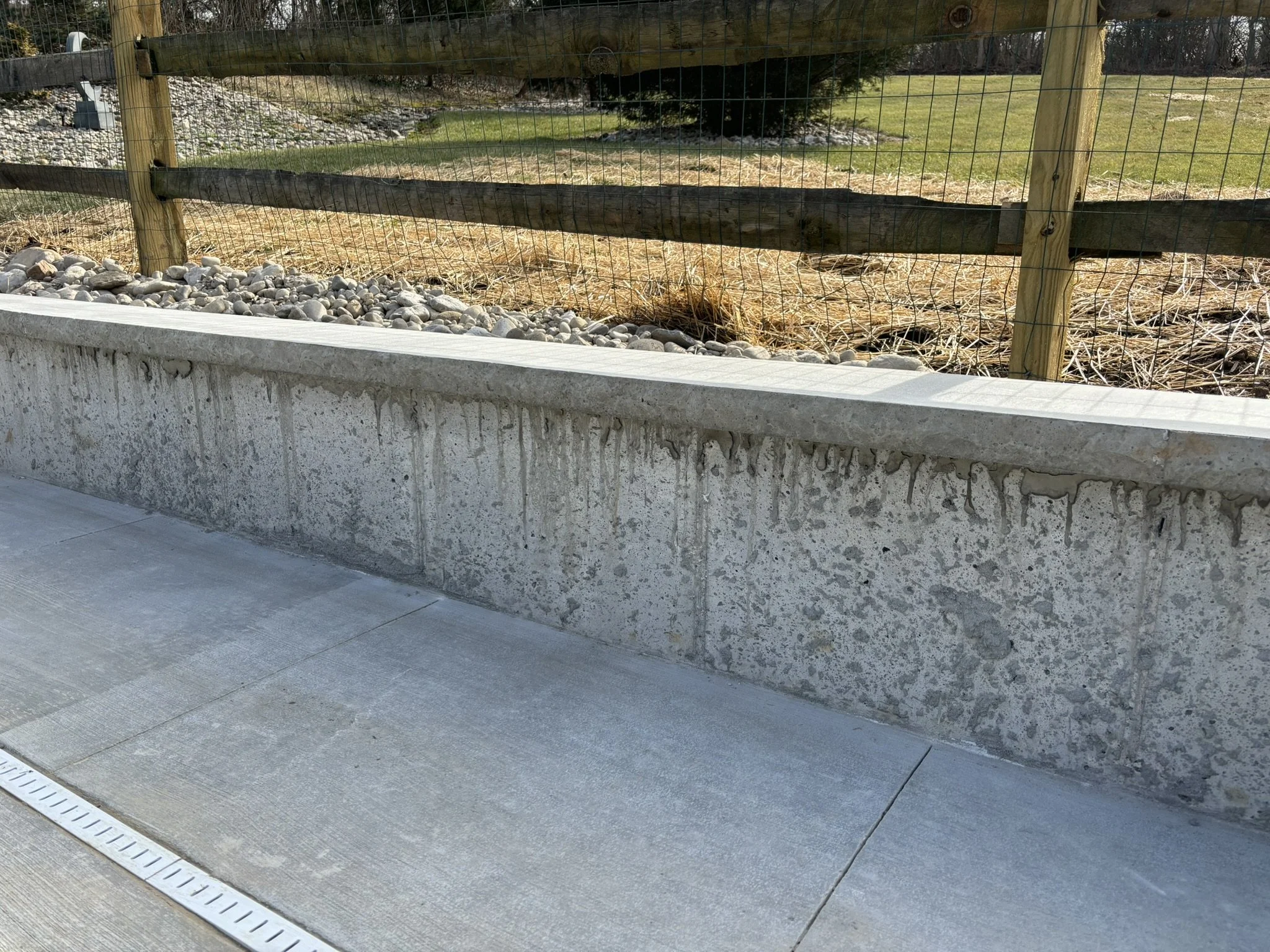 Concrete sidewalk in front of a landscaped area with rocks and a wooden and wire fence, with grass and trees behind.