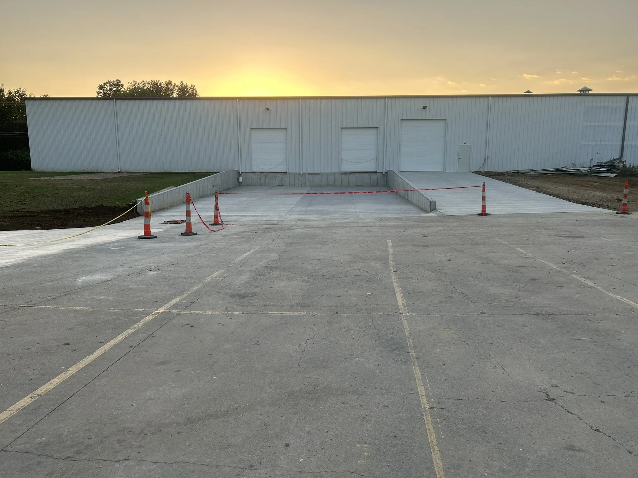 A large white industrial building with three roll-up garage doors in an empty parking lot at sunset. Orange cones and caution tape cordon off part of the area near the building.