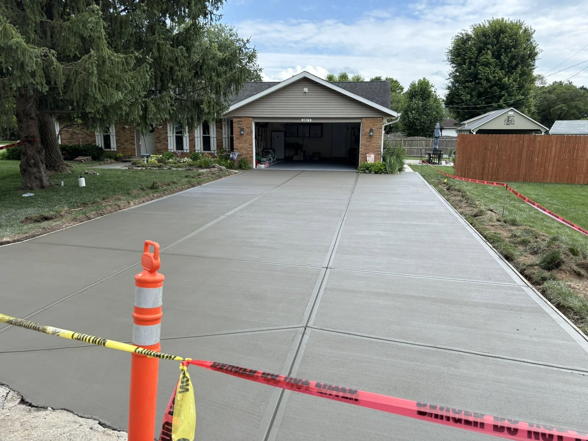 Newly poured concrete driveway in front of a garage with caution tape and traffic cones, surrounded by a residential yard with trees, a wooden fence, and outdoor furniture.