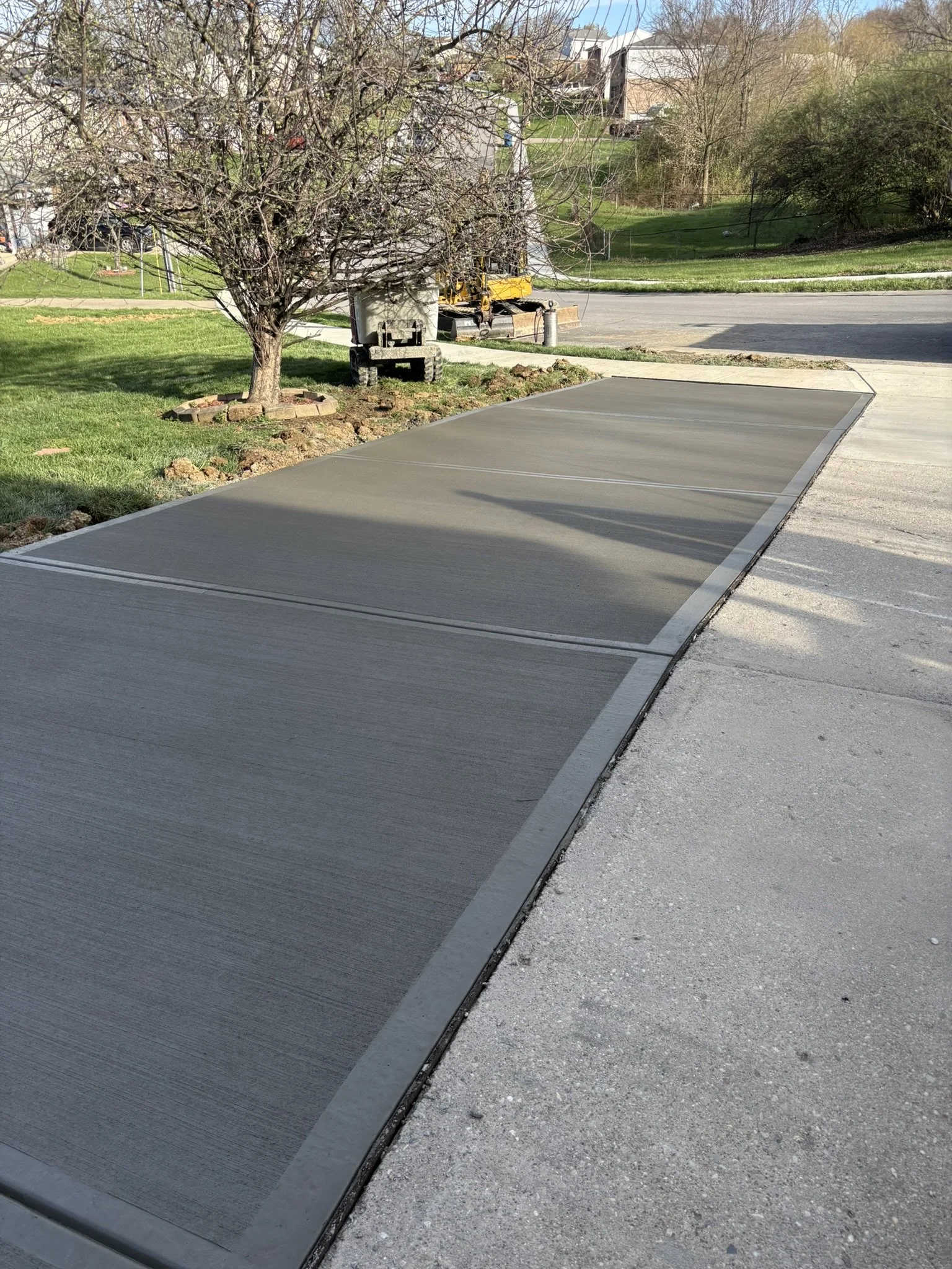 A driveway with a newly poured concrete section, next to a grassy lawn and a leafless tree.