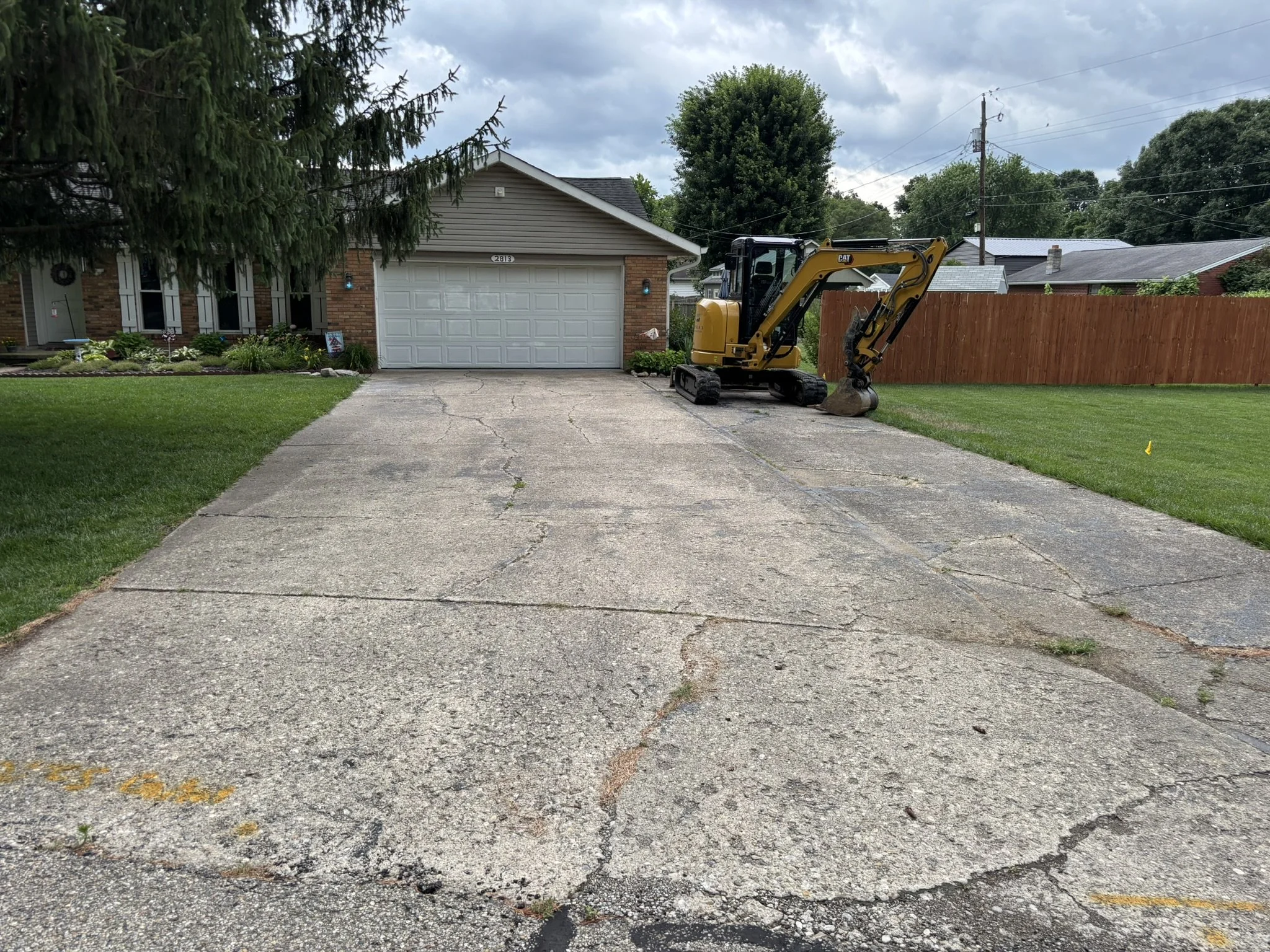 A driveway with cracks, a yellow mini excavator parked on the side, a brick house with a white garage door, and a red wooden fence. Green grass and trees are visible, with a cloudy sky overhead.