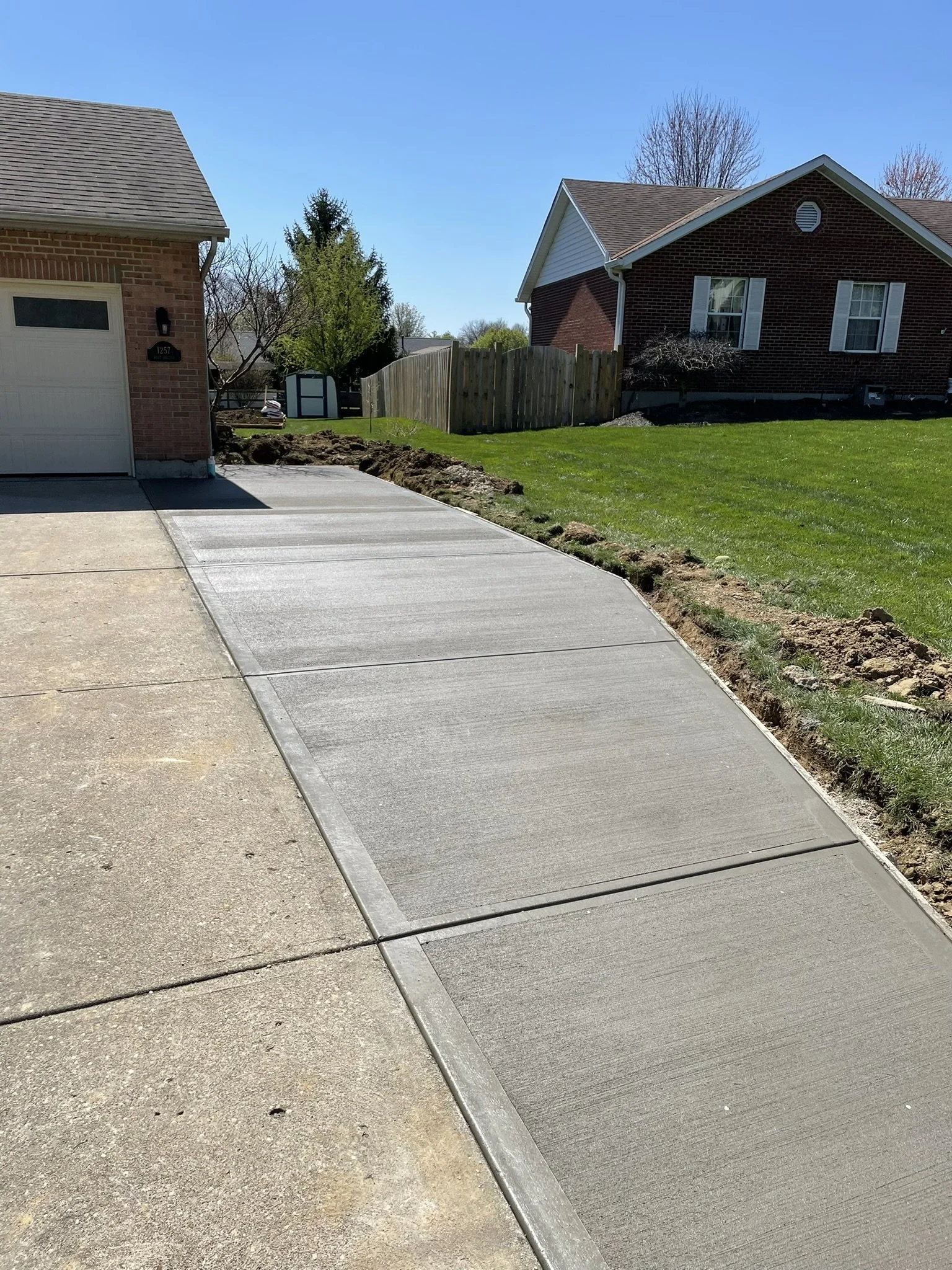 Sidewalk and front yard of a house with a brick facade and an attached garage, green lawn, trees, and clear blue sky.