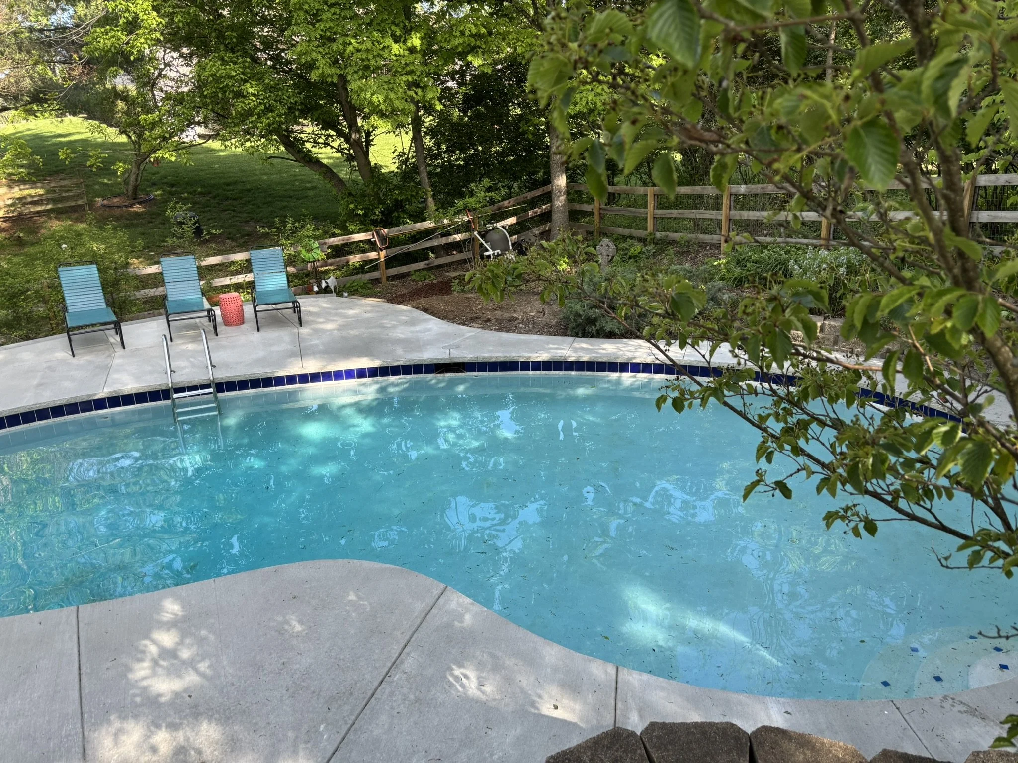 Backyard pool scene with blue lounge chairs, small red table, trees, and garden fencing.