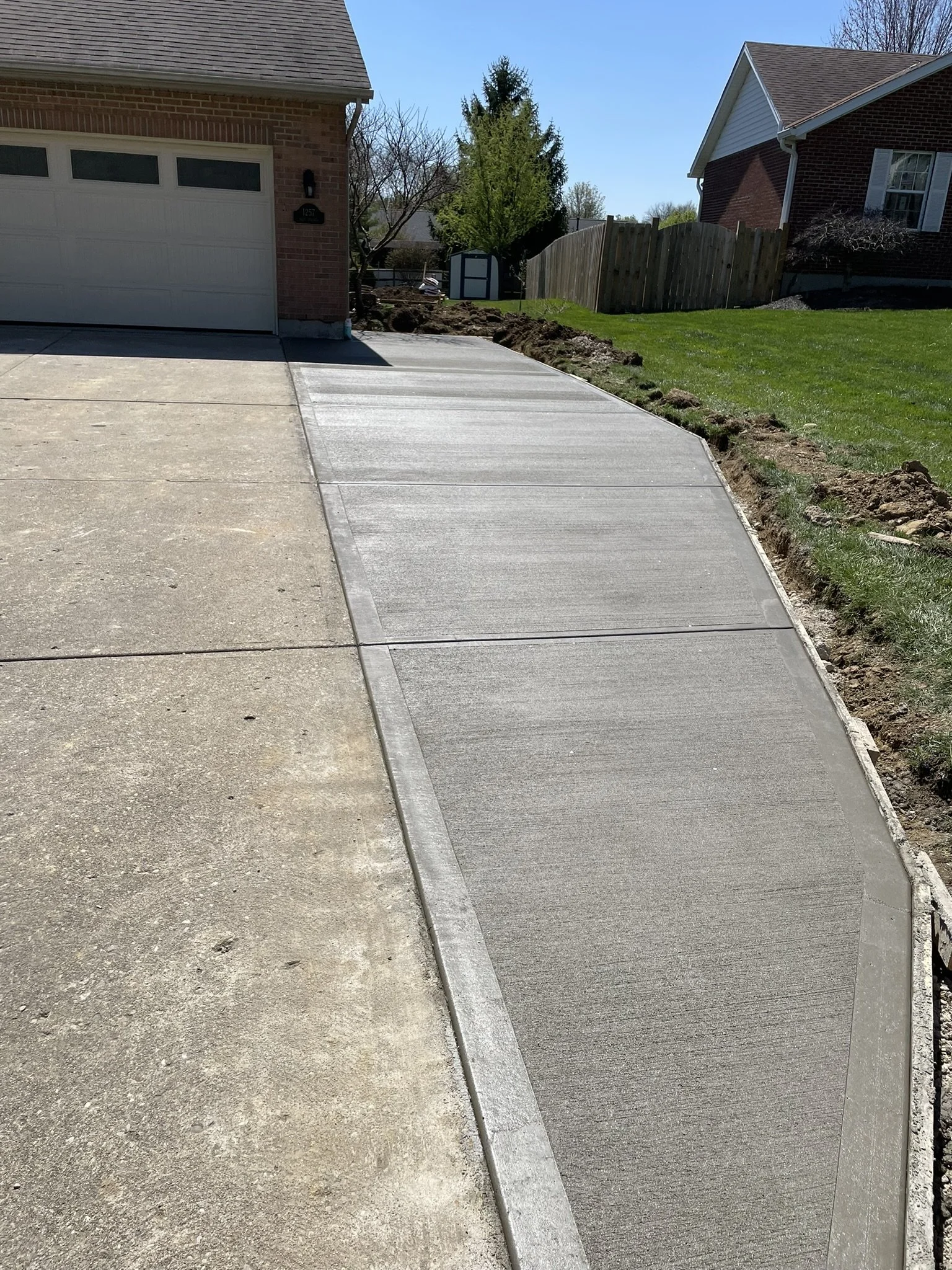 The image shows a residential driveway with a section of concrete and a section of stone-paved concrete, next to a house with a brick facade and a garage door. There is a fenced backyard with some trees and a shed, and a grassy lawn area.