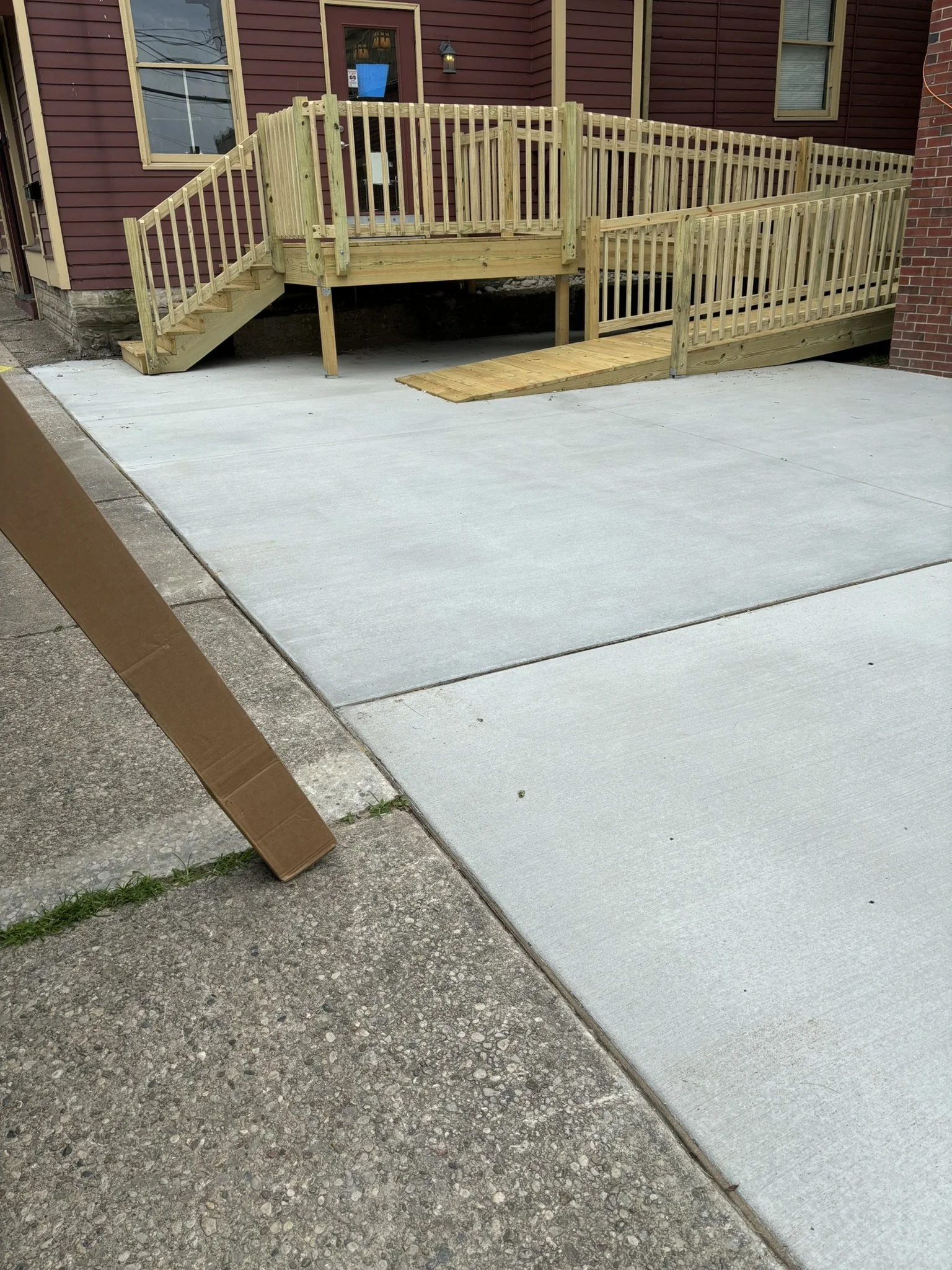 New wooden ramp and stairs installed outside a house with a maroon exterior, brick foundation, and multiple windows, leading from the sidewalk to the house entrance.