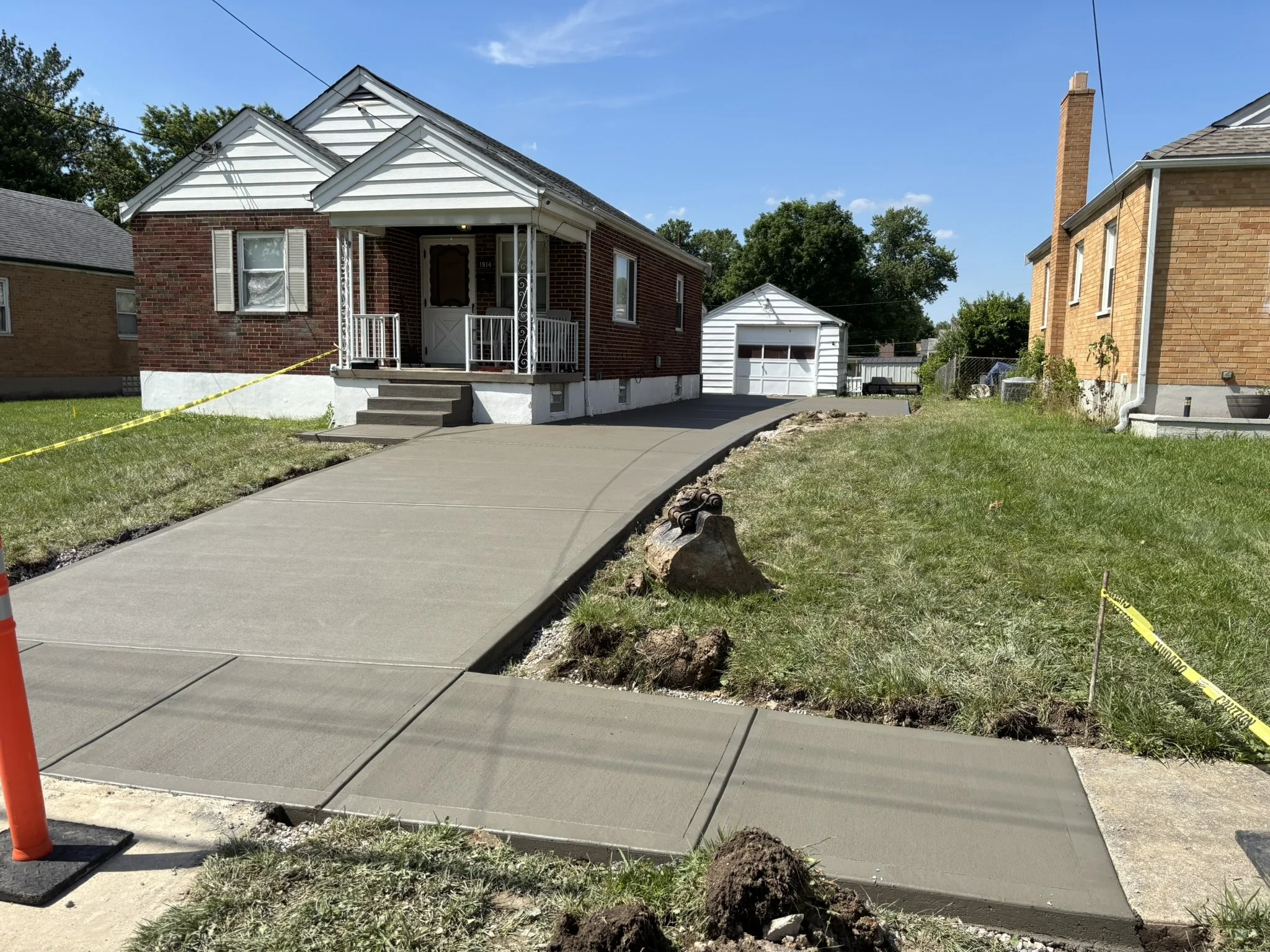 A newly poured concrete sidewalk with sections separated by expansion joints, adjacent to a grassy yard. A construction cone and yellow caution tape are present, indicating ongoing work. Two brick houses are visible, one with a small screened porch a