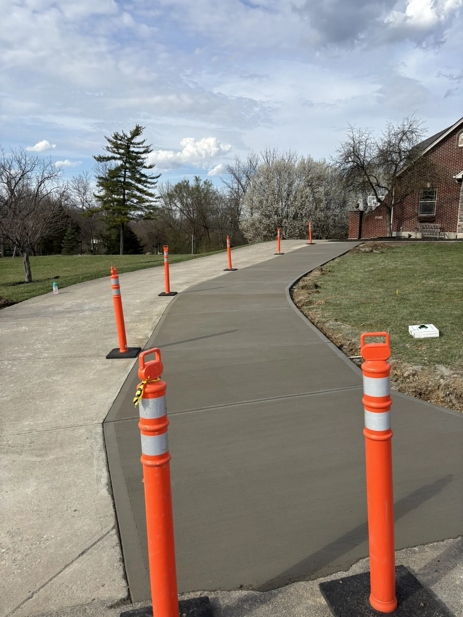 Newly poured concrete sidewalk with orange traffic cones and a small orange safety barrier, leading up to a brick house with trees and a cloudy sky in the background.