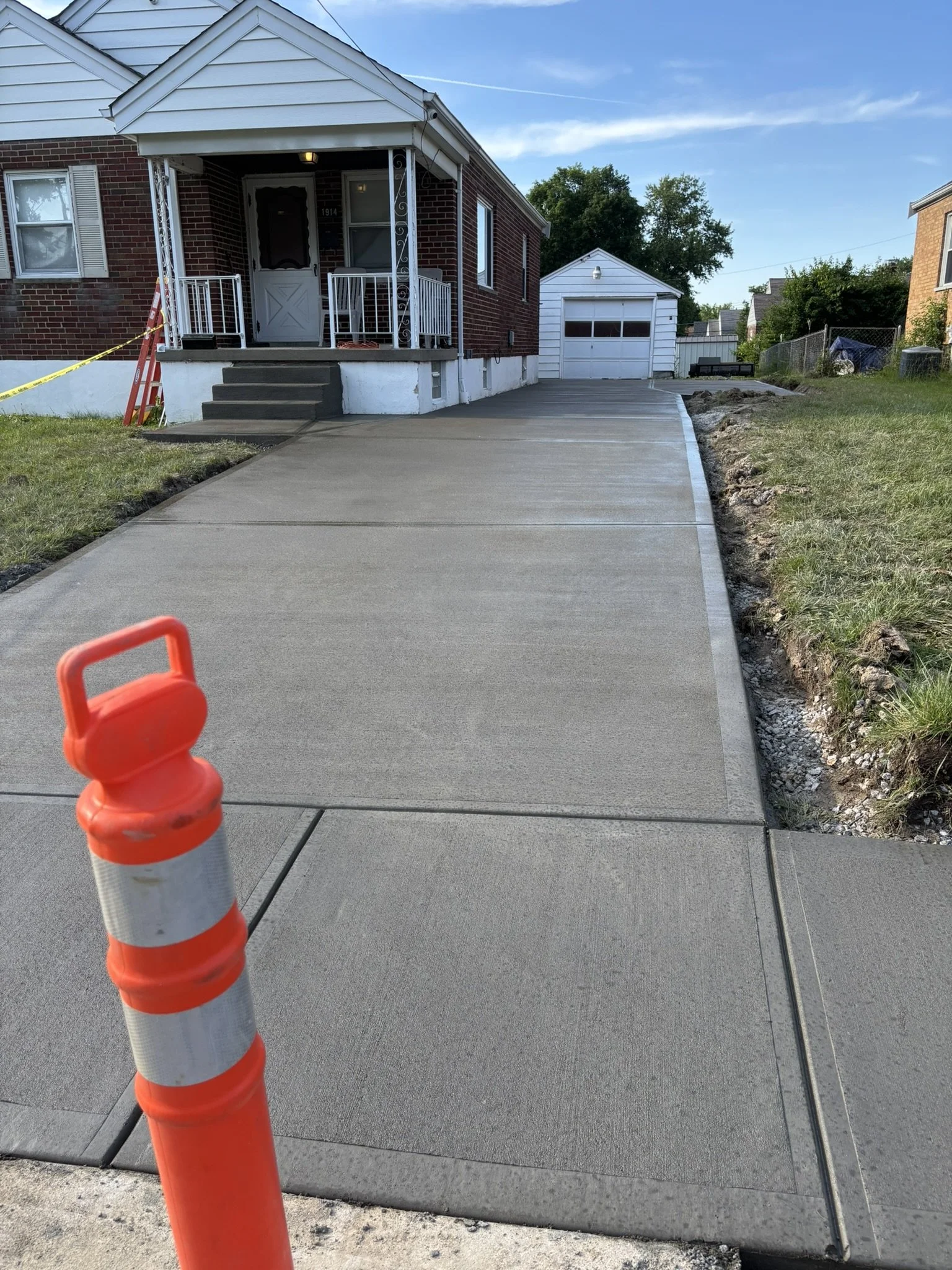 Newly poured concrete sidewalk leading to a house with brick exterior, white siding, front porch, and steps. Construction cone in foreground and a new garage in the background.