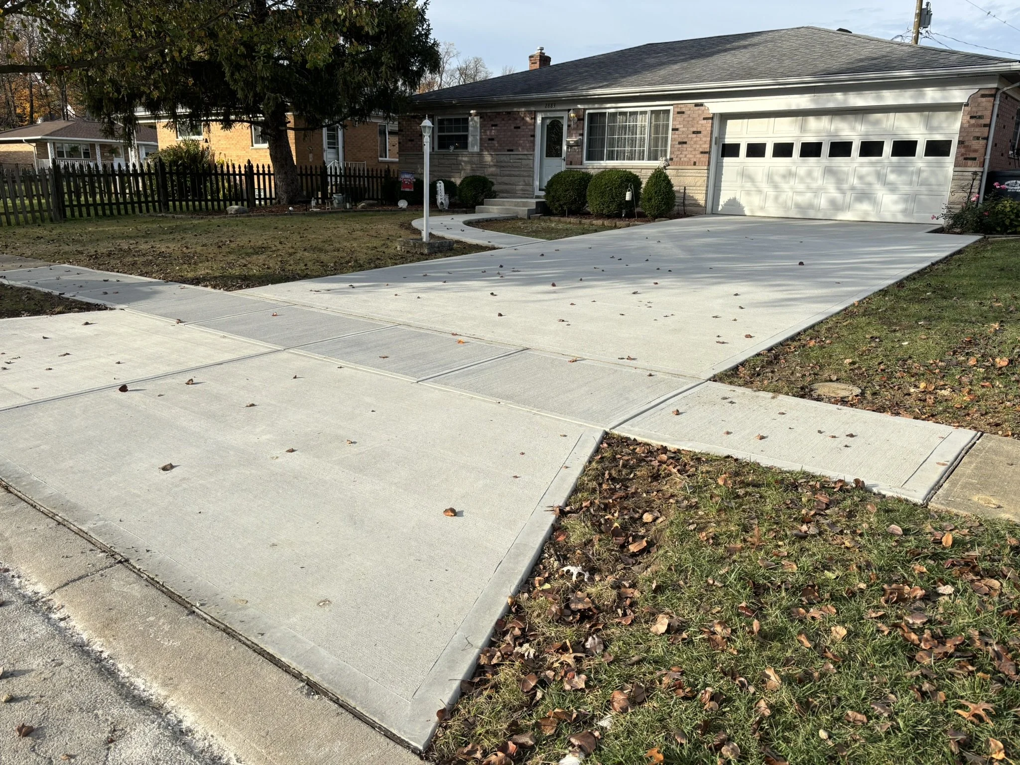 A suburban house with a well-maintained front yard, concrete driveway, and a white garage door. A large tree shades part of the yard, and a black wooden fence lines the property. Fall leaves are scattered on the driveway and lawn.