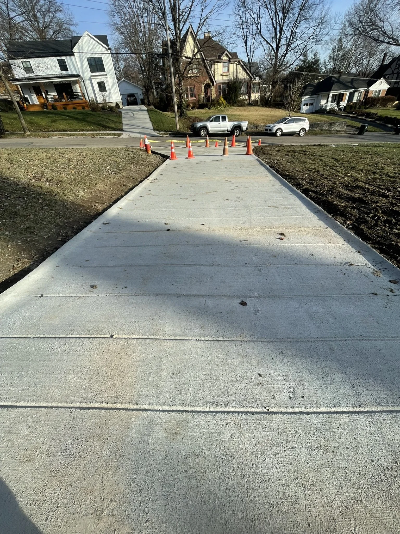 Freshly poured concrete sidewalk with highway cones blocking the walkway, residential houses in the background on a clear day.