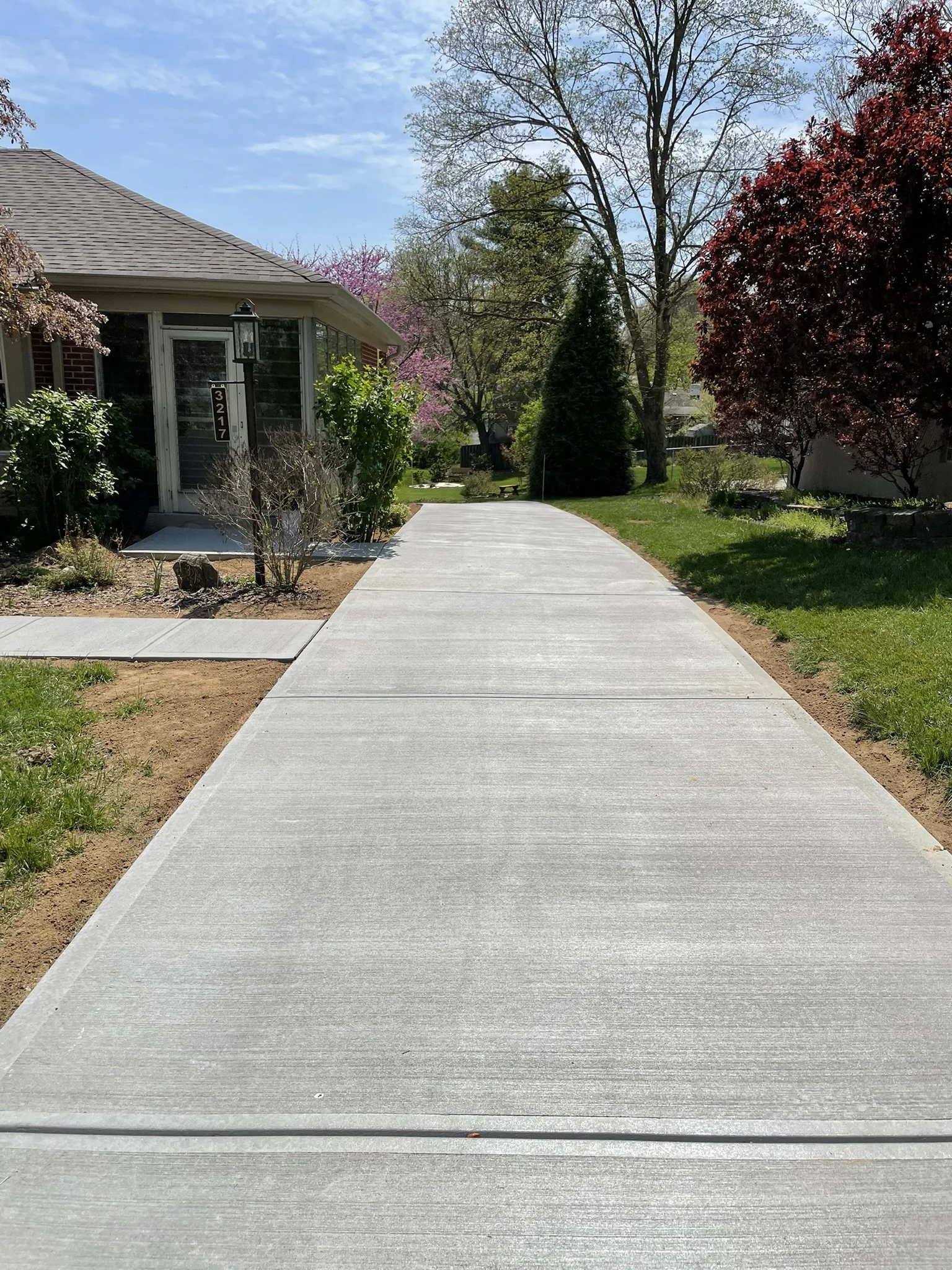 Concrete sidewalk leading to a house with a garden and trees on a sunny day.