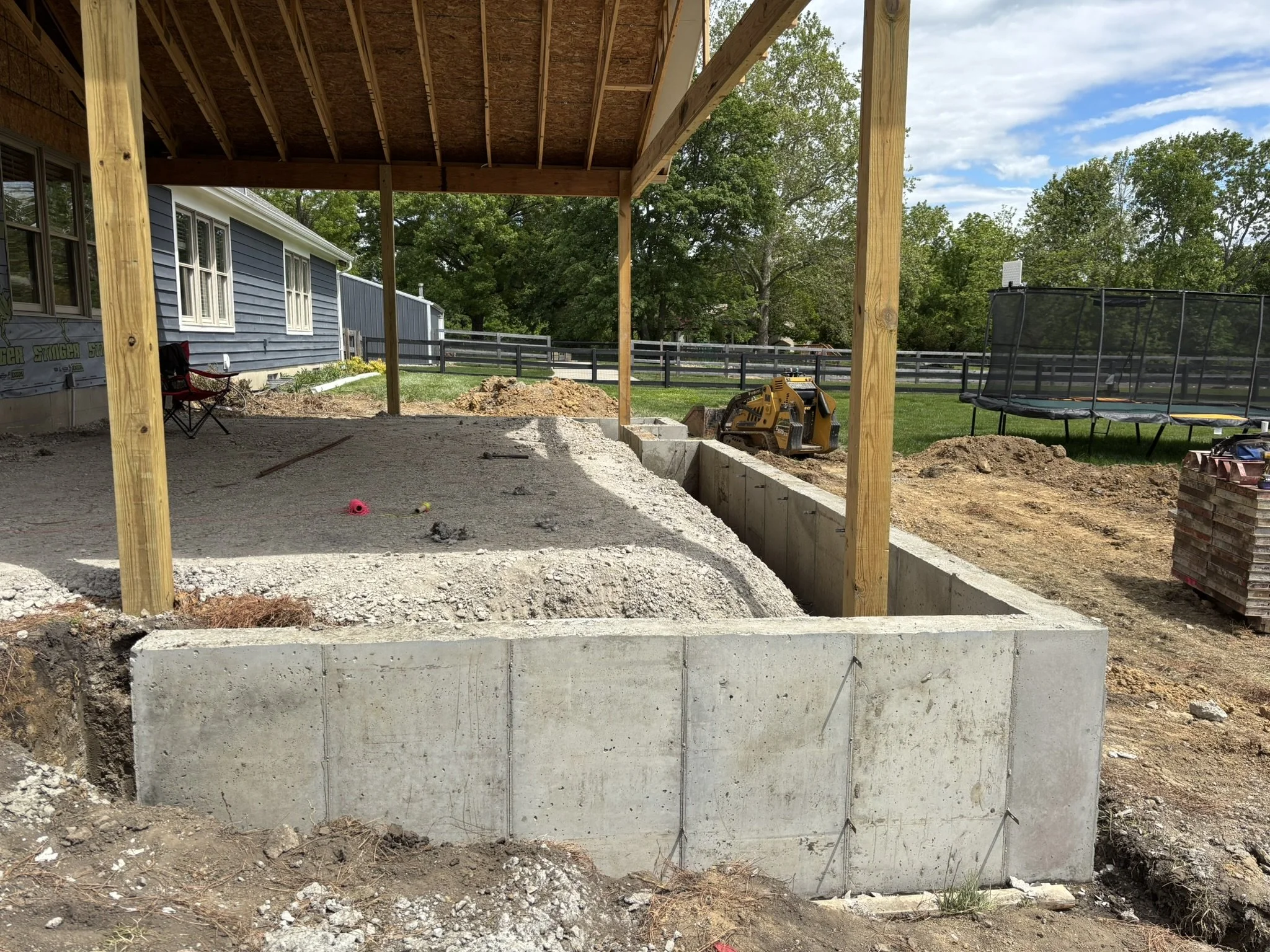 A backyard under construction with a new patio foundation, wooden posts for a deck, and construction equipment on the grass. There are trees and a blue house with white trim in the background under a partly cloudy sky.