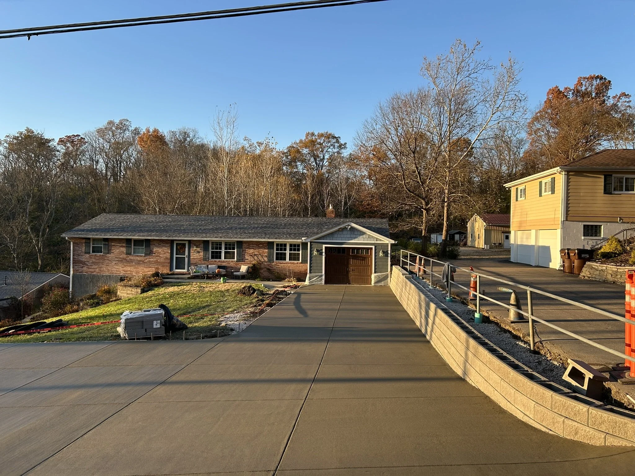 Residential backyard with a house, a garage, a patio with outdoor furniture, a partially finished driveway, and trees in the background under a clear blue sky.