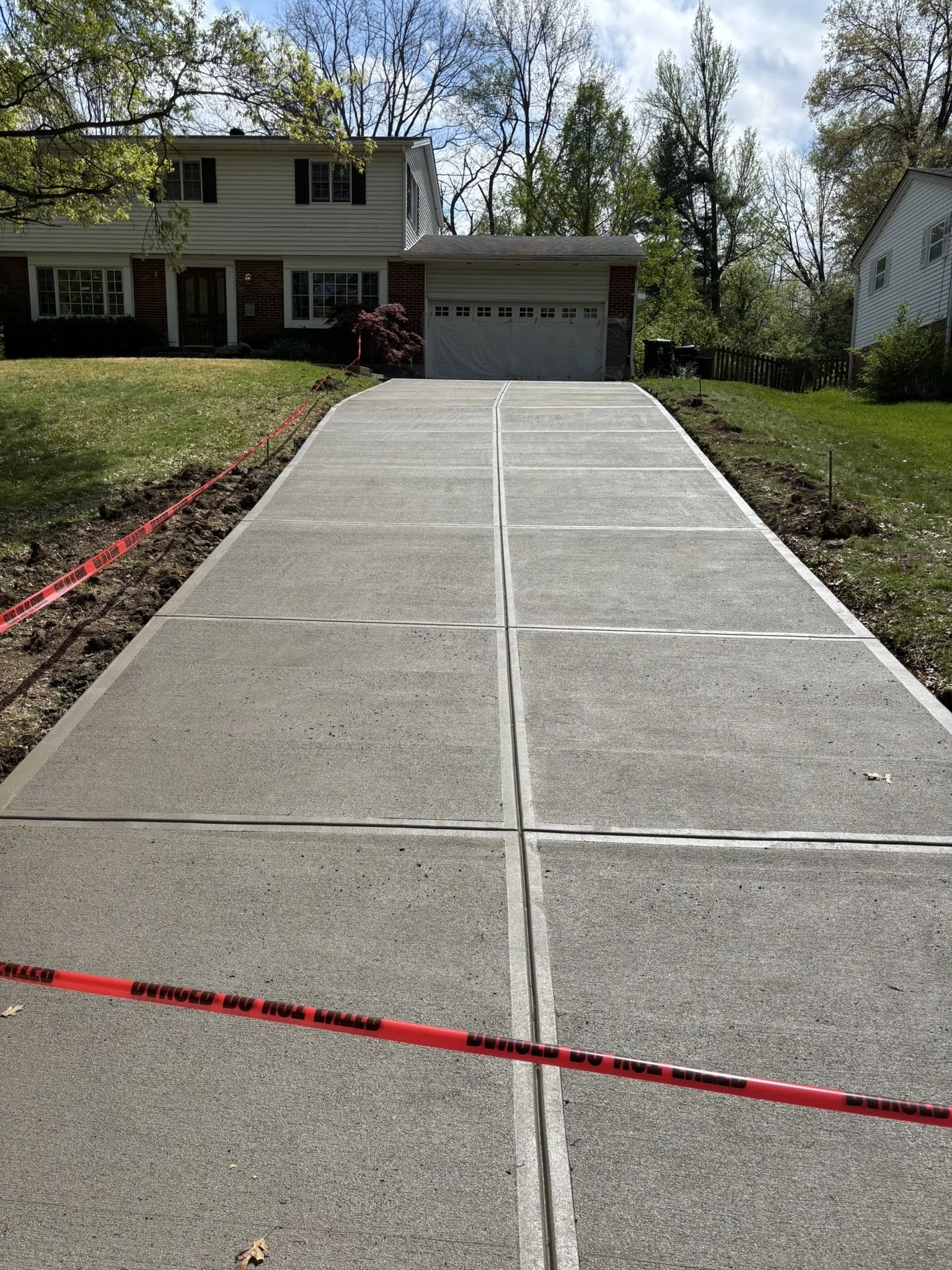 Newly poured concrete driveway with safety warning tape in front of a suburban house, with trees and neighboring houses visible behind.