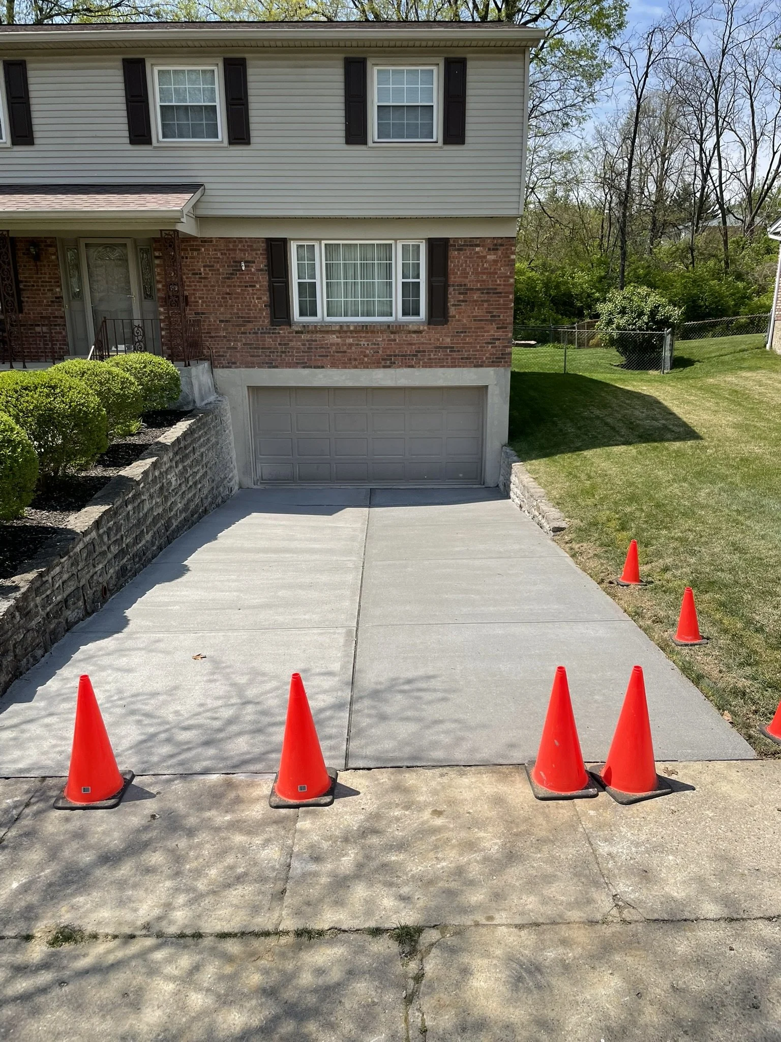 Newly poured concrete driveway with orange traffic cones lined up along its edges for curing or construction purposes, adjacent to a residential house with a brick and siding exterior, a double garage, and a lawn.