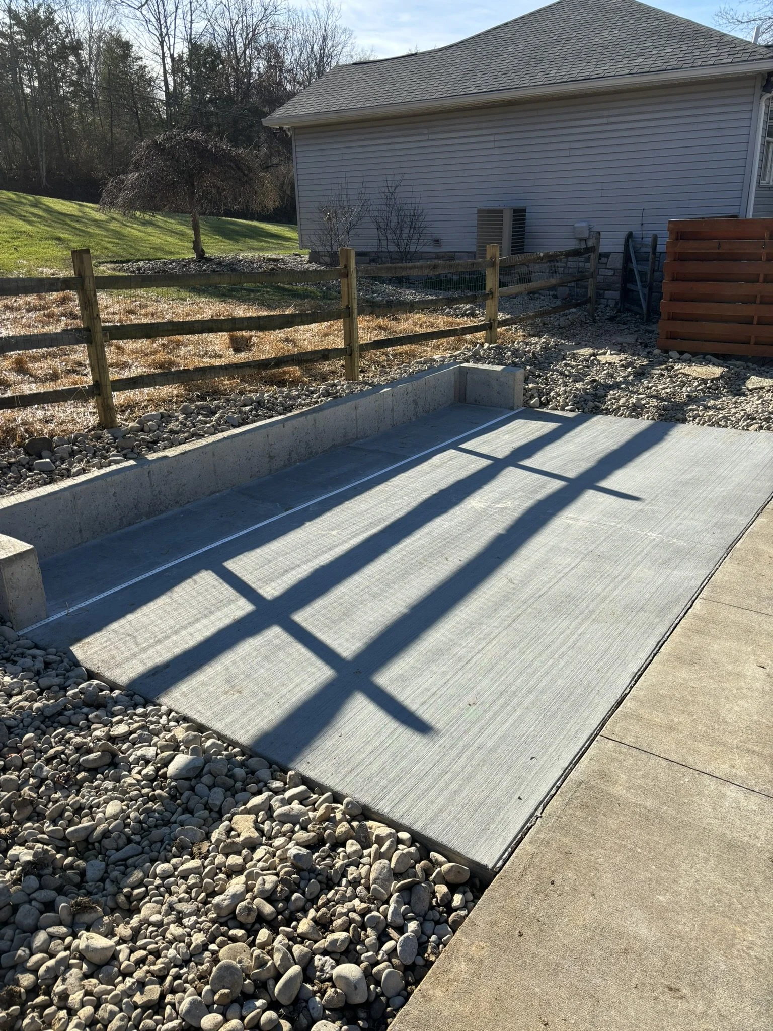 Empty garden bed frame on a concrete patio with a gravel border, in a backyard with grass, trees, and a house wall visible in the background.