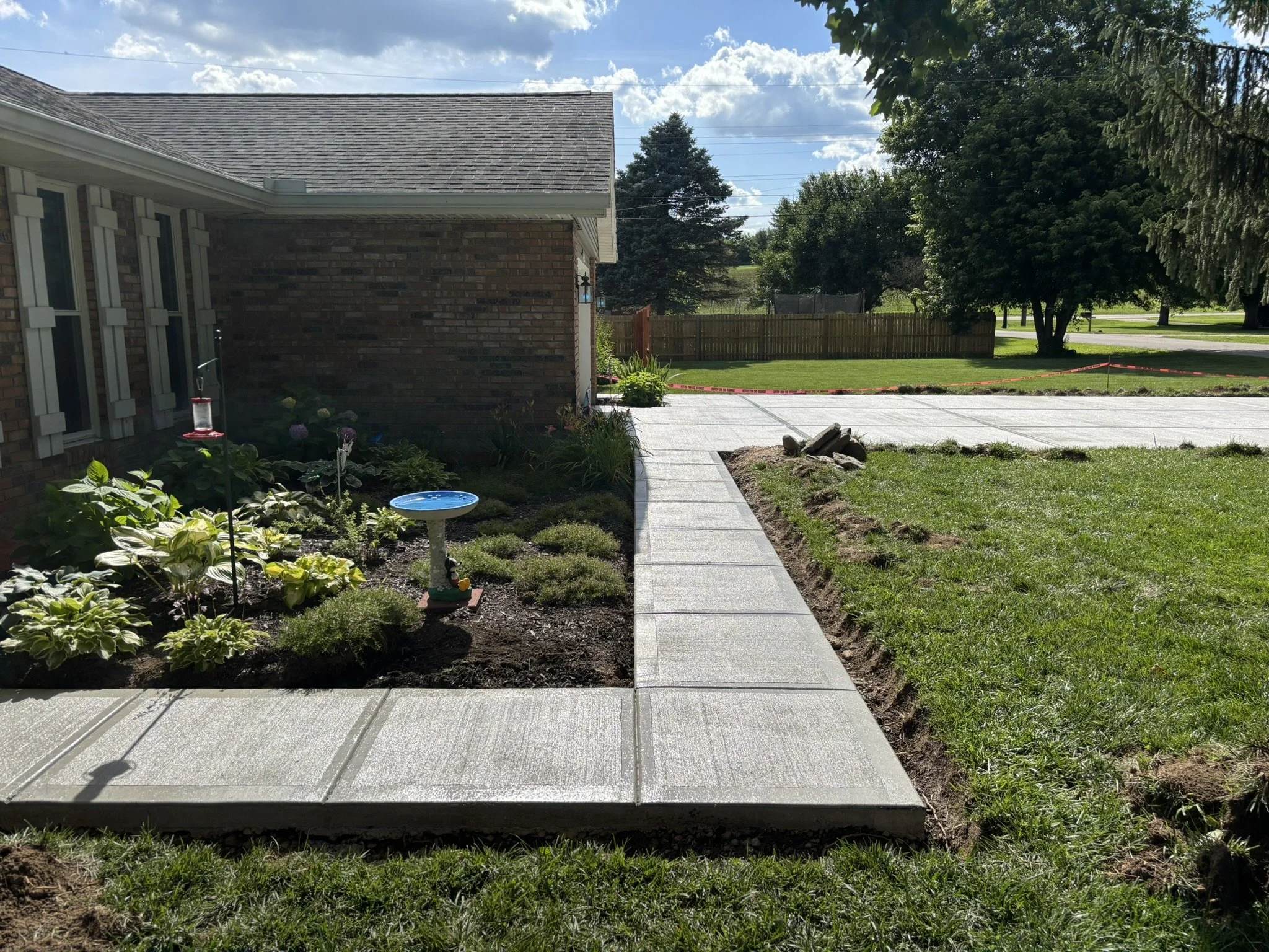 A backyard with a brick house on the left, a paved walkway in the middle, a grassy lawn to the right, a wooden fence, trees, and a partly cloudy sky.