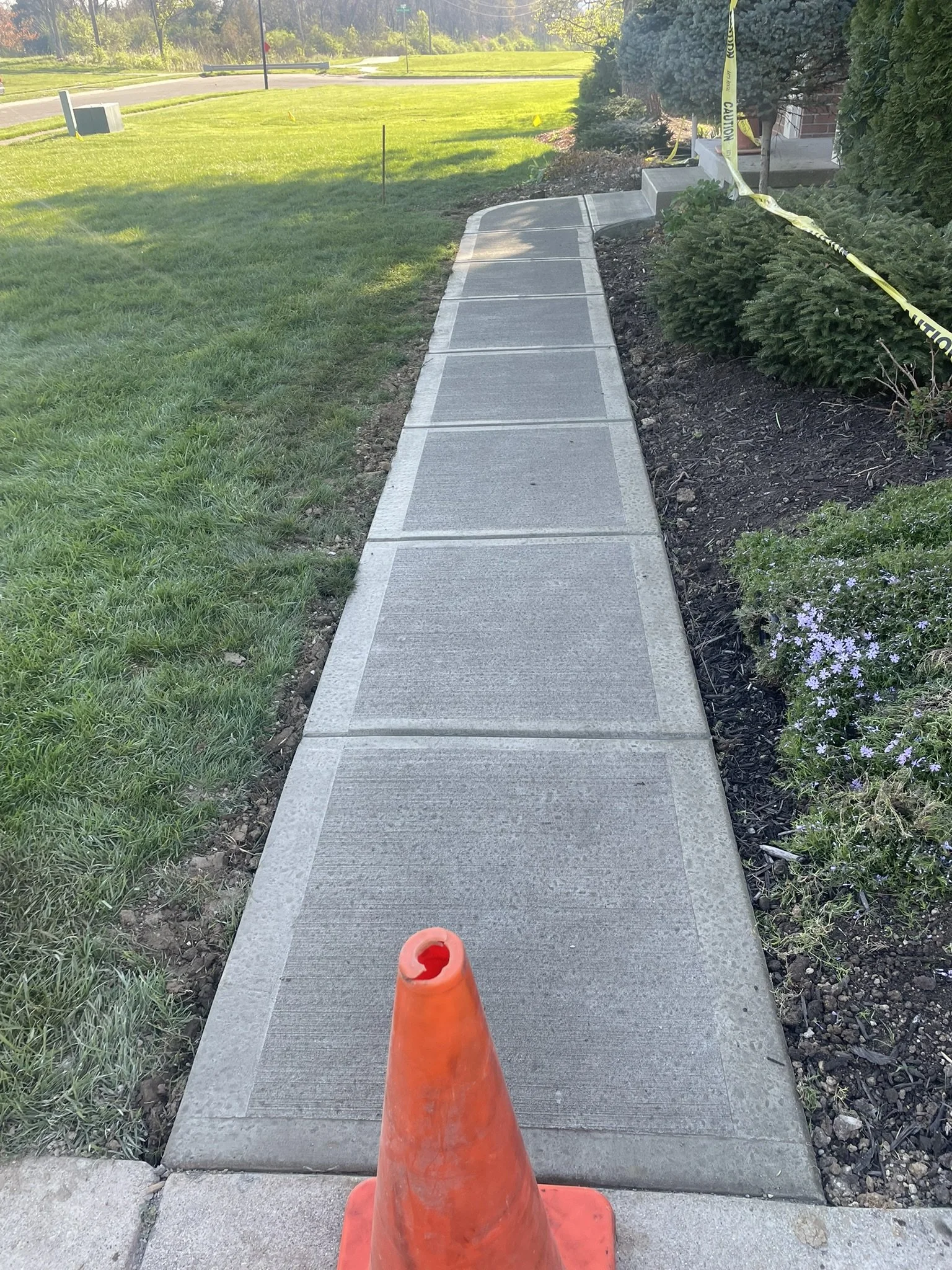 A new concrete sidewalk with a construction orange cone at the beginning, surrounded by grass and landscaping, with caution tape on bushes and a street visible in the background.