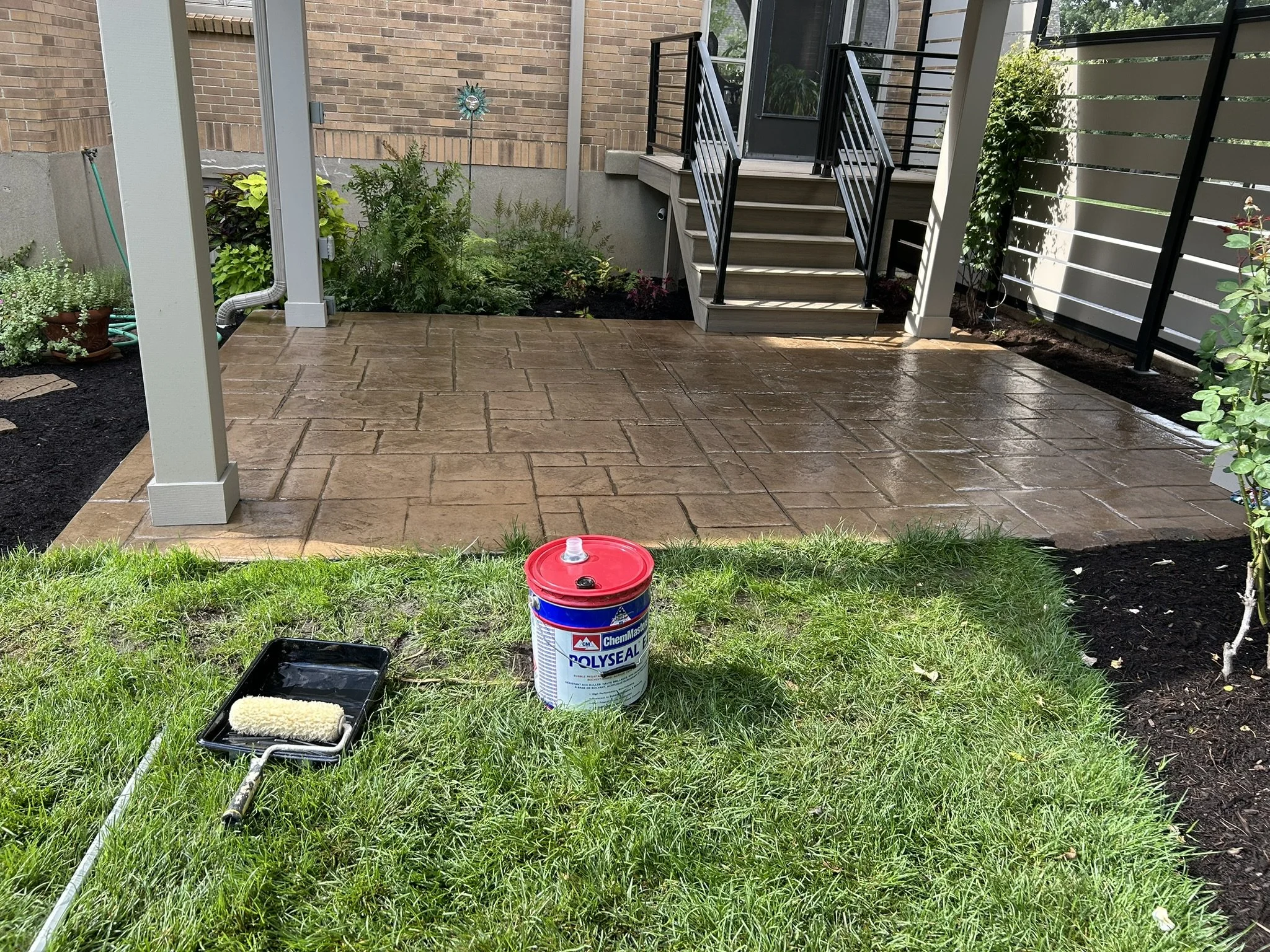 A backyard patio with a newly laid stamped concrete surface, a small garden bed with green plants, a staircase leading to a deck, and painting supplies including a paint roller, tray, and a bucket of Polysealer concrete sealer, with a grassy area in 