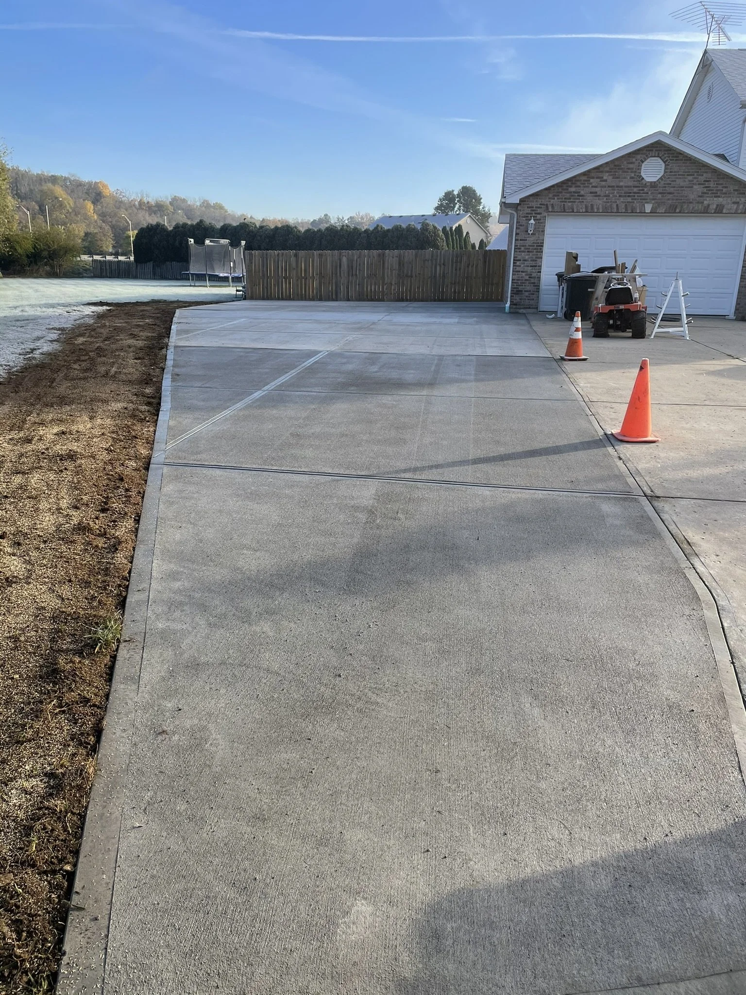 A newly poured concrete driveway outside a suburban house, with safety cones and construction tools nearby, and a wooden fence at the back.