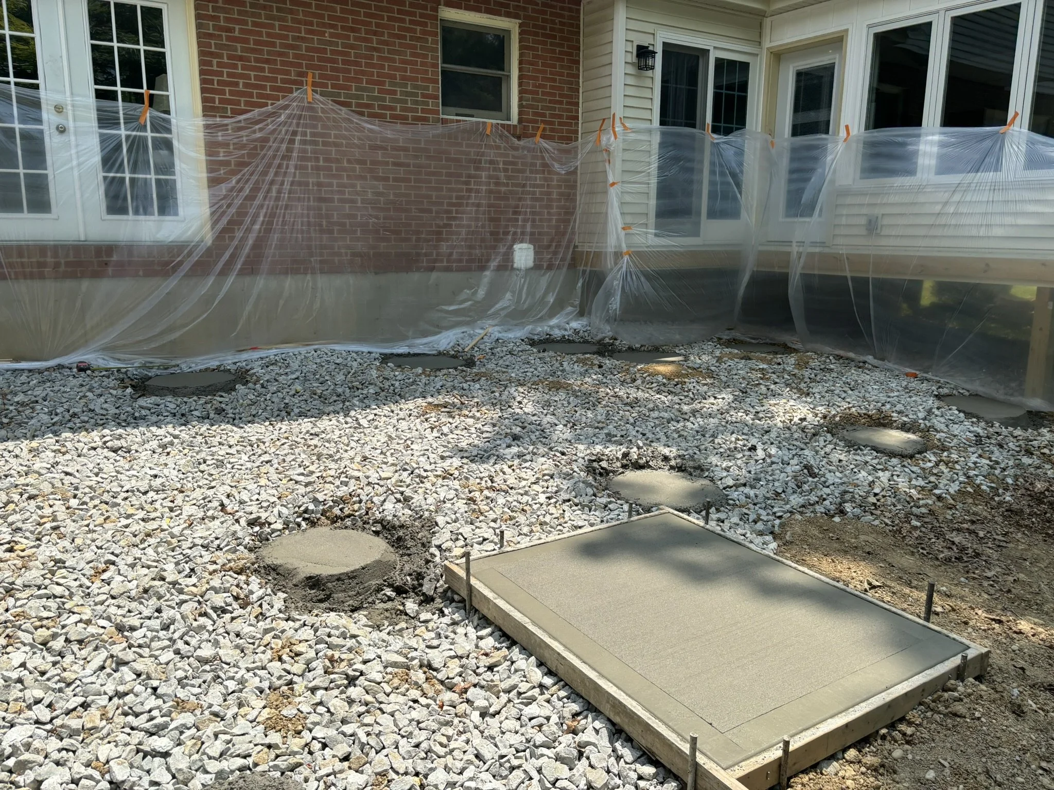 Backyard with a brick wall house, sliding glass doors, and a gravel yard. Clear plastic sheeting attached to the house with orange clips, covering the bottom portion of the wall.