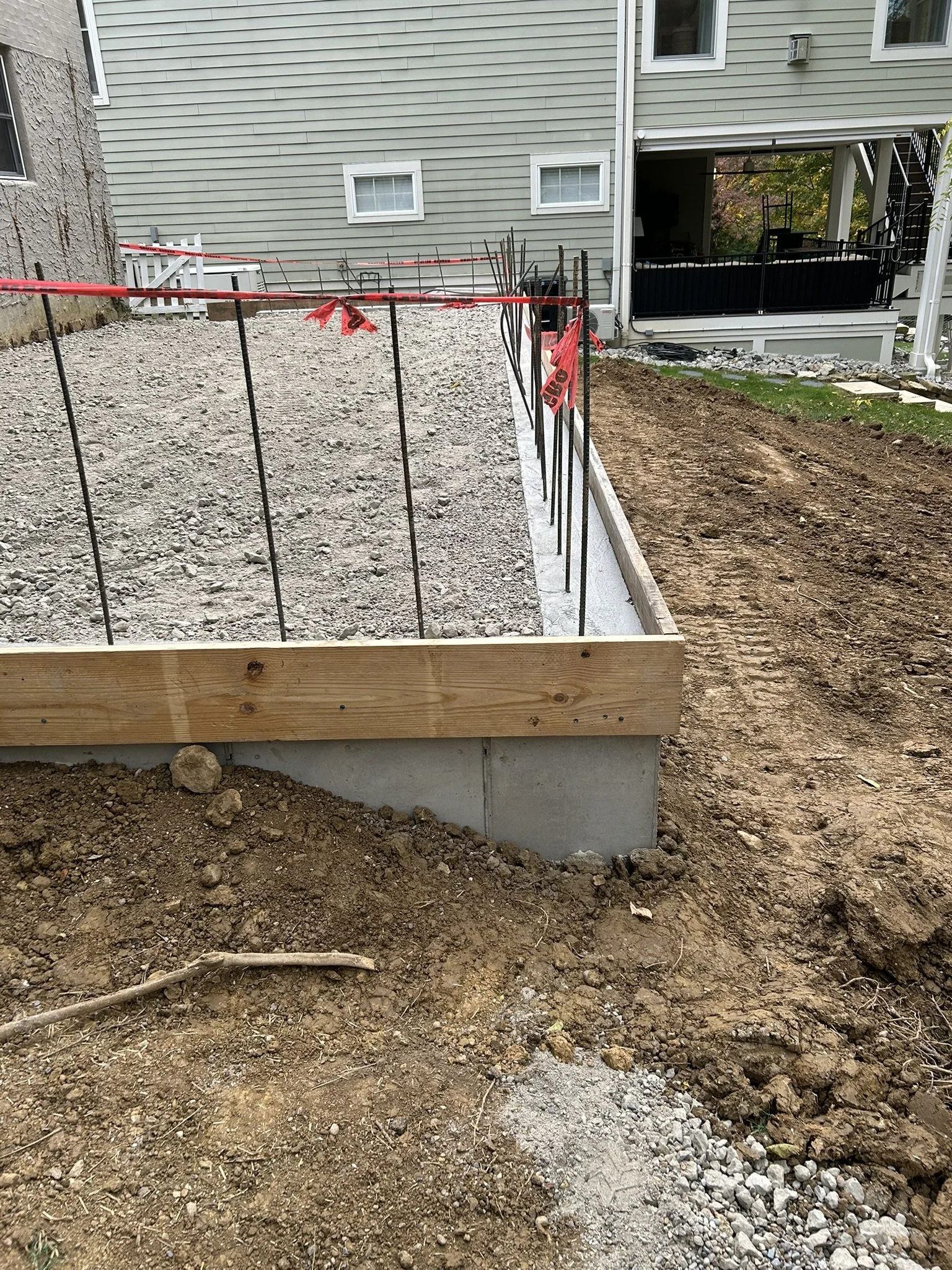 Construction site with dug up dirt and gravel, wooden and metal framing, and house with green siding in background.