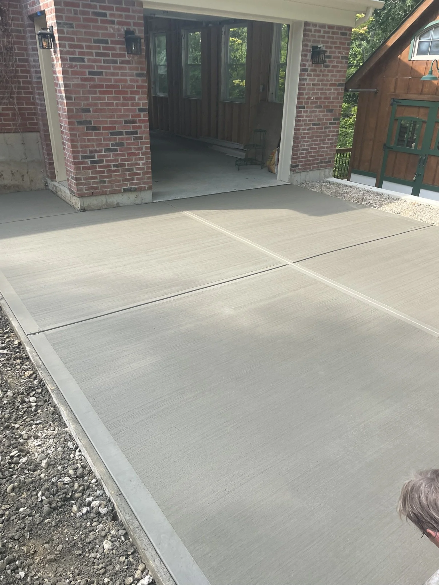 Freshly poured concrete in front of a brick house with a garage opening and a small child with light-colored hair in the corner.