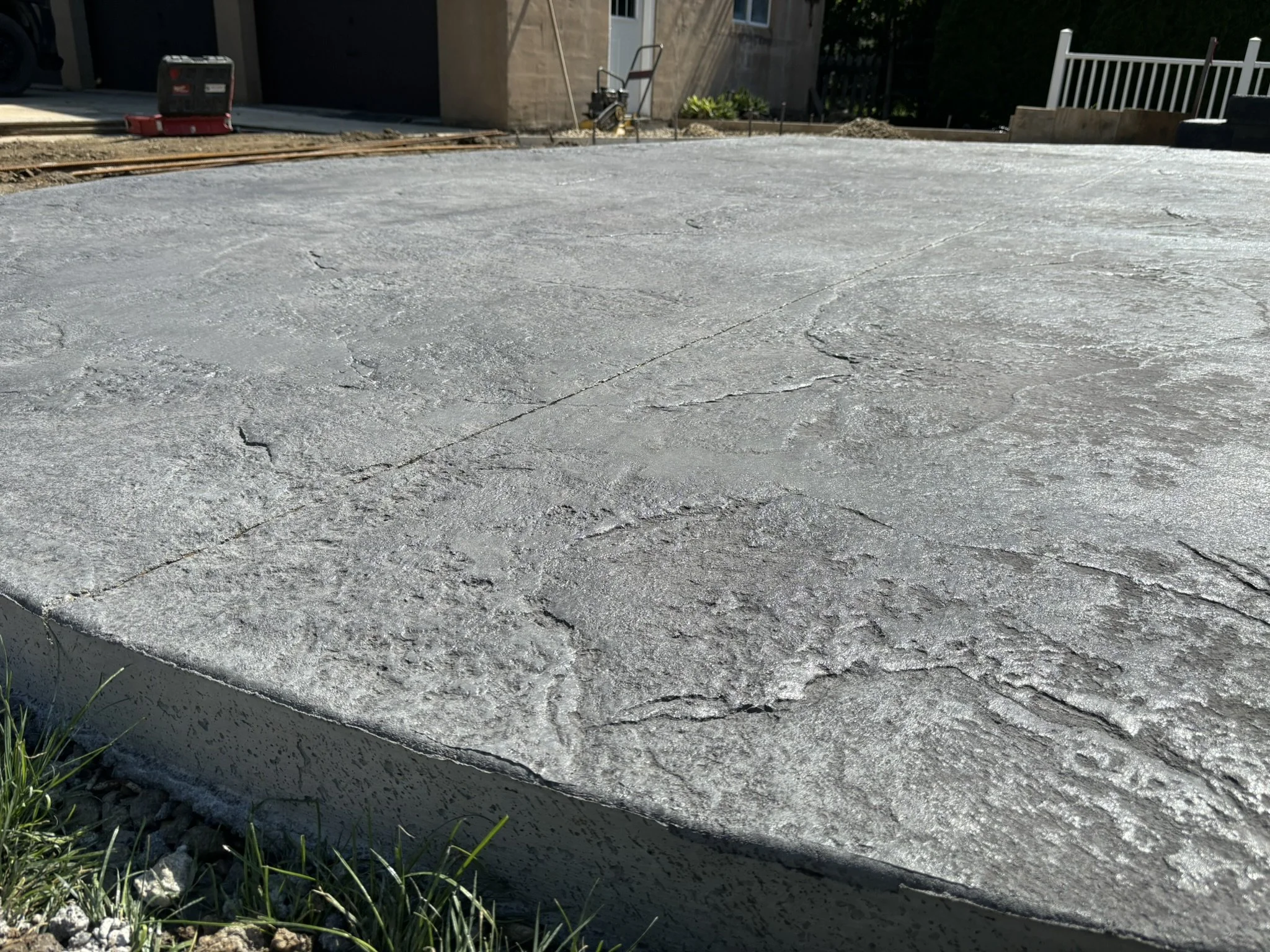 Close-up of a freshly poured concrete foundation at a construction site with a curb and grass in the foreground, and a building with construction tools in the background.