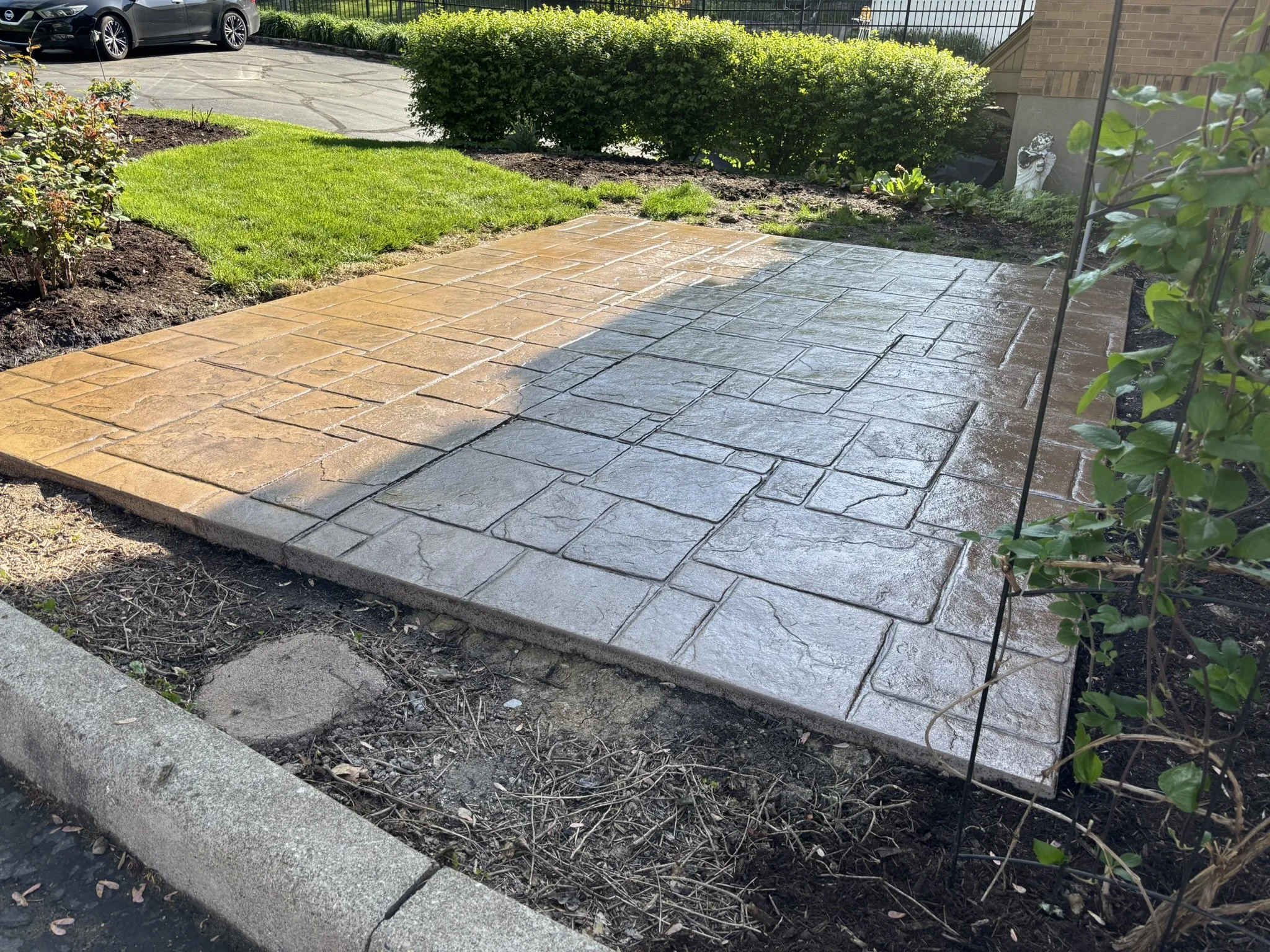 Newly paved stone patio with wet surface, adjacent to a garden bed with green grass and bushes, located next to a parking lot and a brick building.