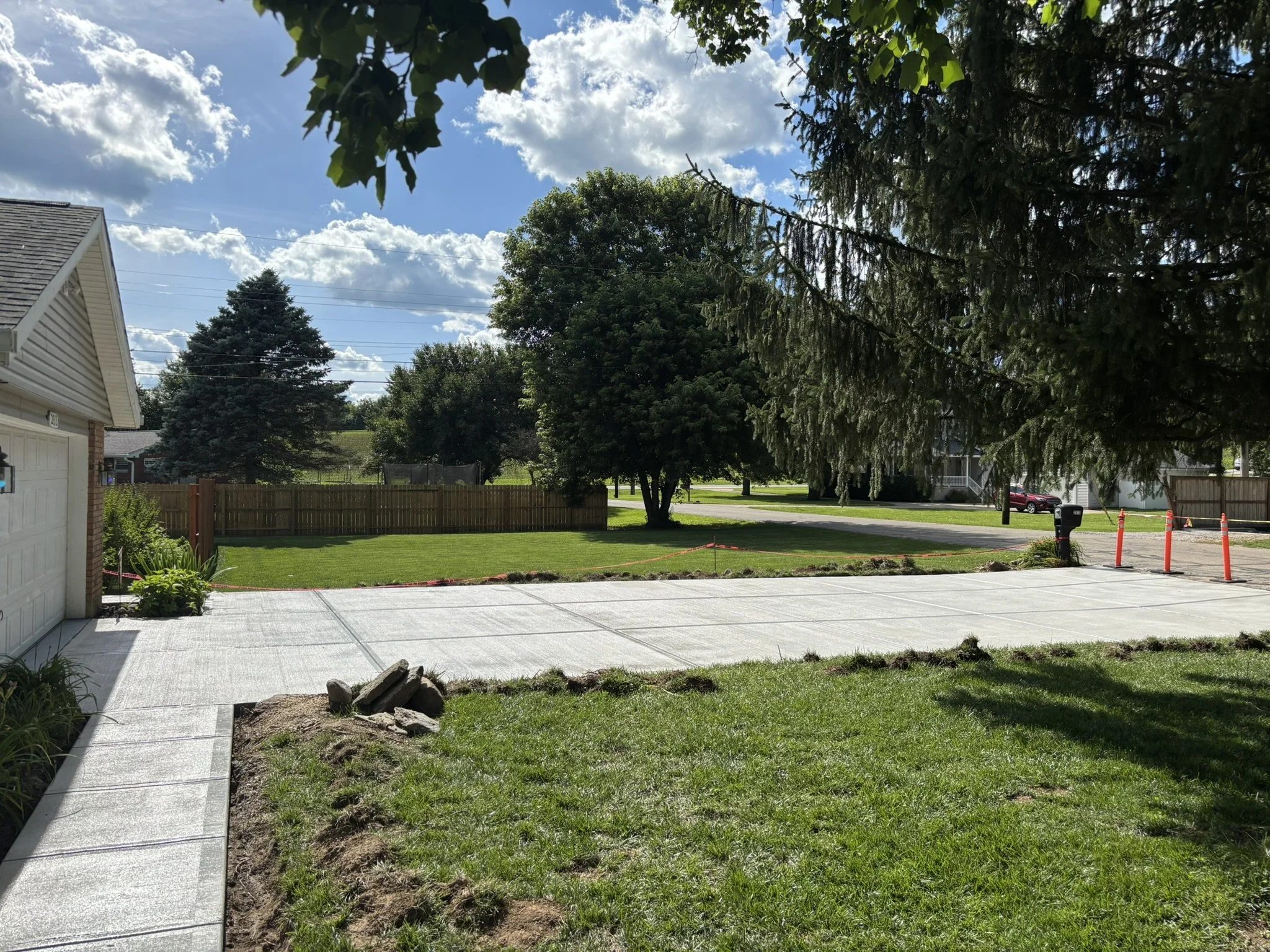 A backyard scene with a concrete patio, green lawn, trees, a wooden fence, and houses in the background under a partly cloudy sky.