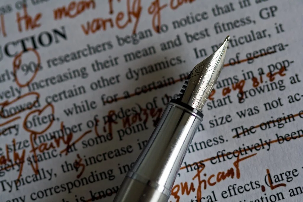 Close-up of a silver fountain pen resting on a page with black printed text, with handwritten annotations and underlines in orange ink.