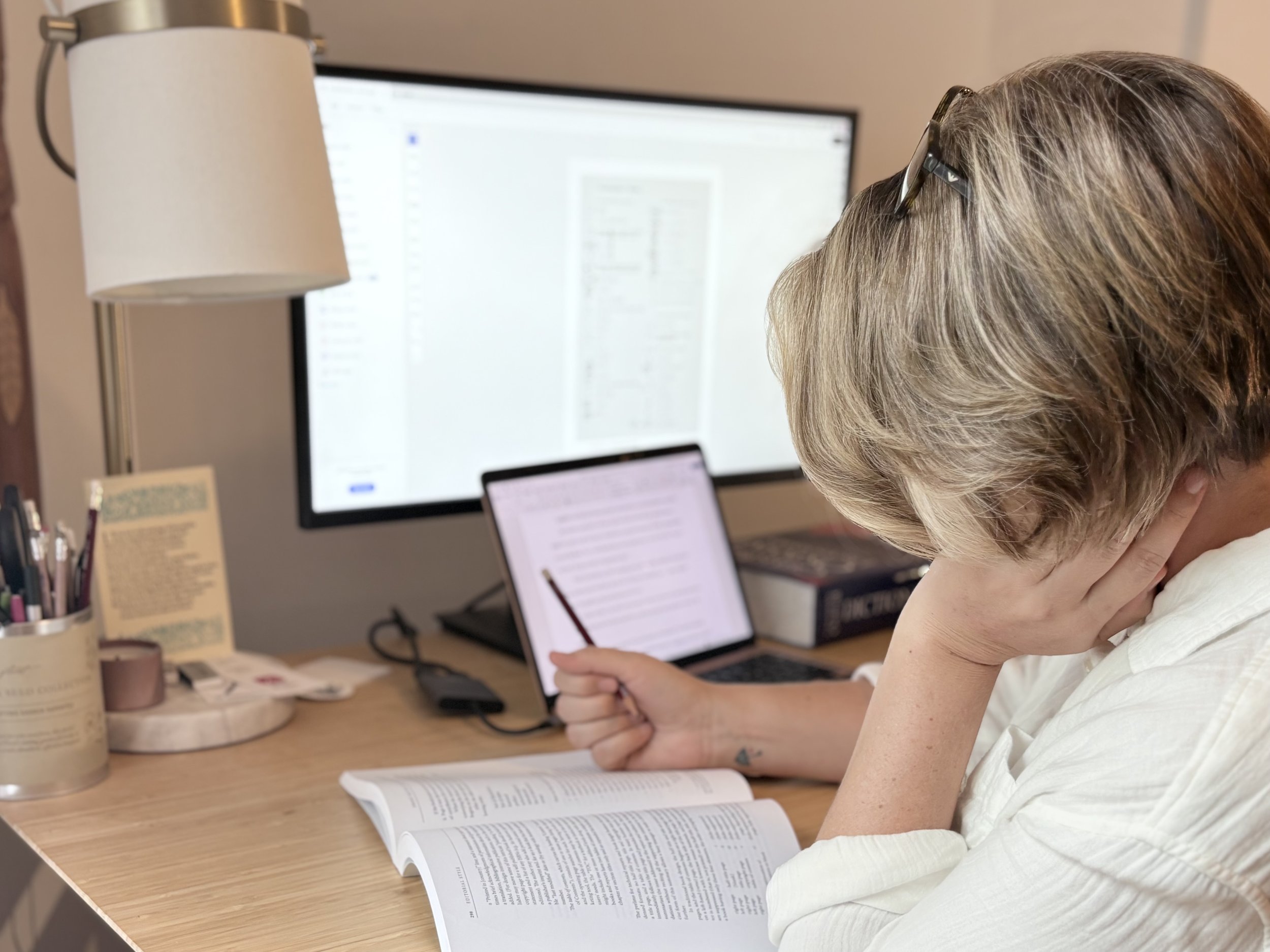 Fiction editor Allison Richard,  with short blond hair and glasses, is reading a book at her desk with a large computer monitor in the background. The desk has a laptop, documents, and writing utensils, with a lamp and a container of pens visible.