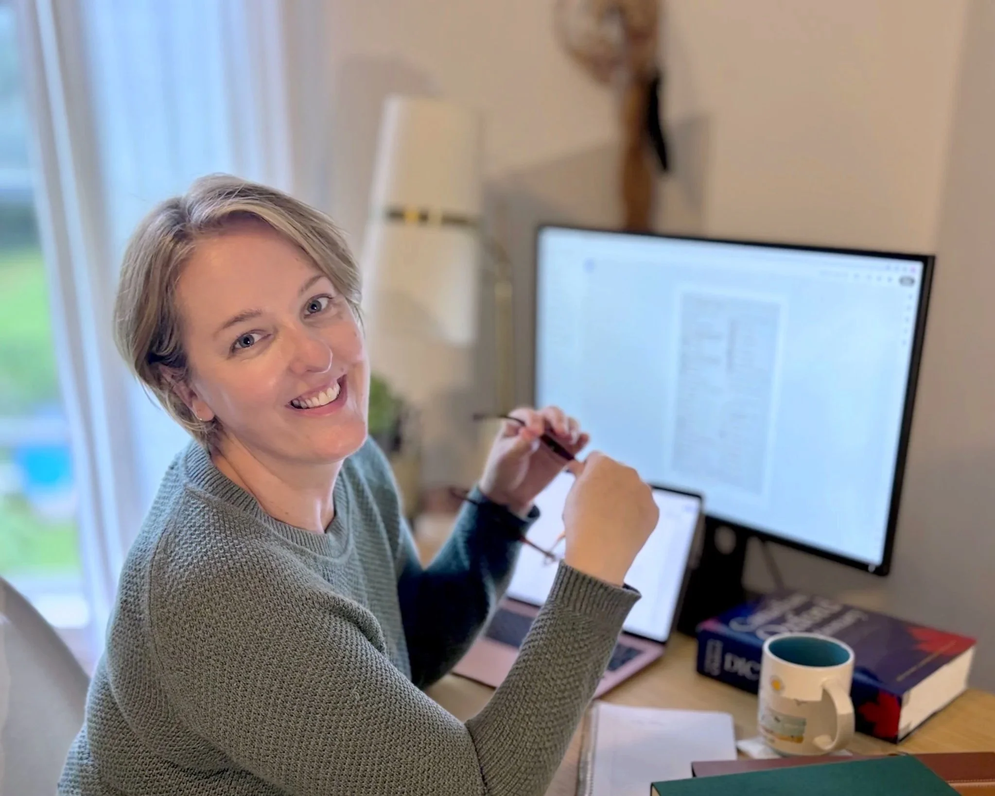 Professionally trained editor Allison Richard wearing a green sweater,  sitting at a desk, smiling, with a computer monitor in front of her. Behind her, a lamp and a window with curtains are visible, along with a mug and some notebooks on the desk.
