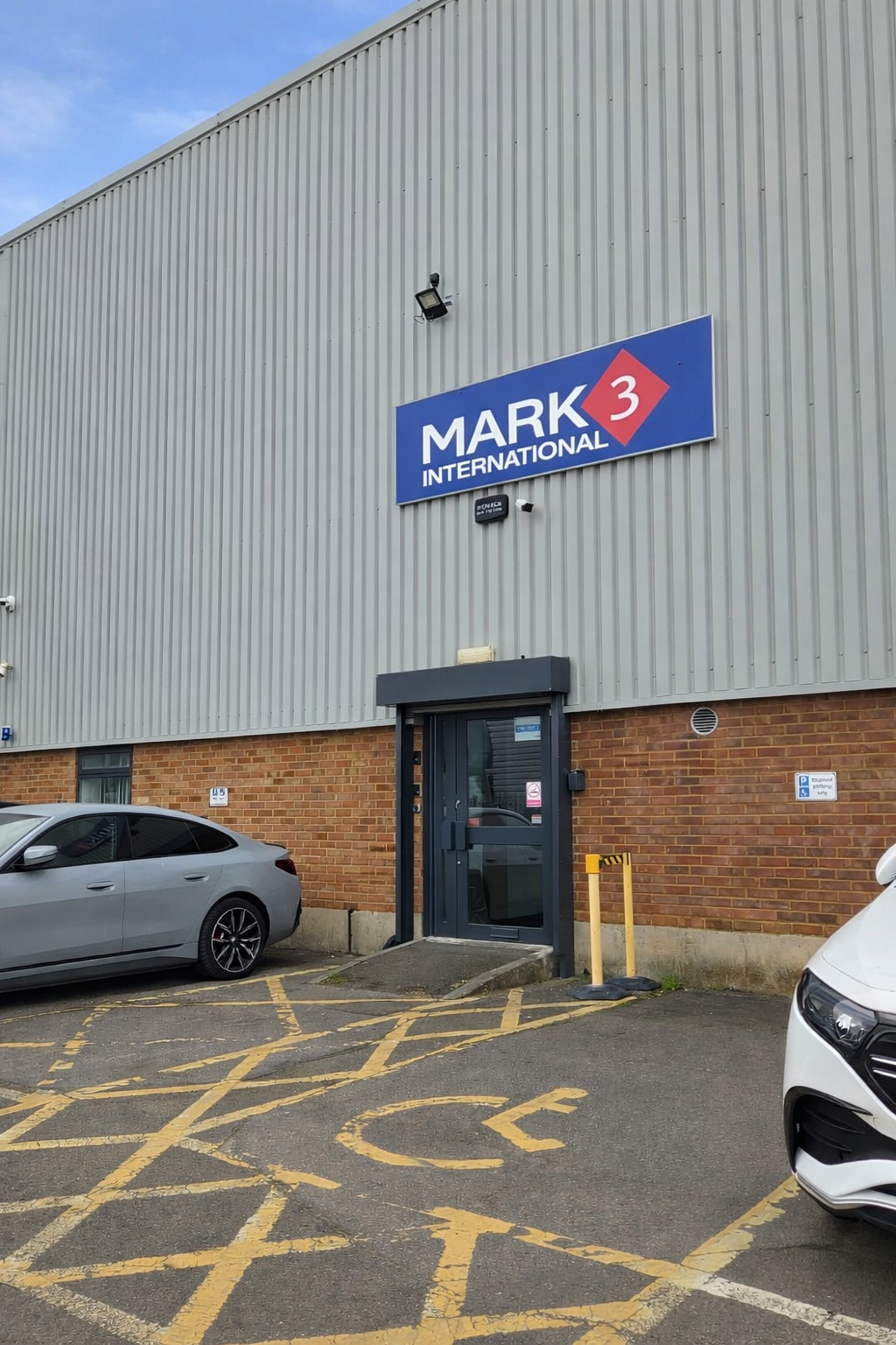 Exterior view of a building with a sign that says 'MARK 3 INTERNATIONAL.' The building has a gray corrugated metal upper section and a brick lower section. There are parking spaces in front, including a designated disabled parking space with a yellow wheelchair symbol.