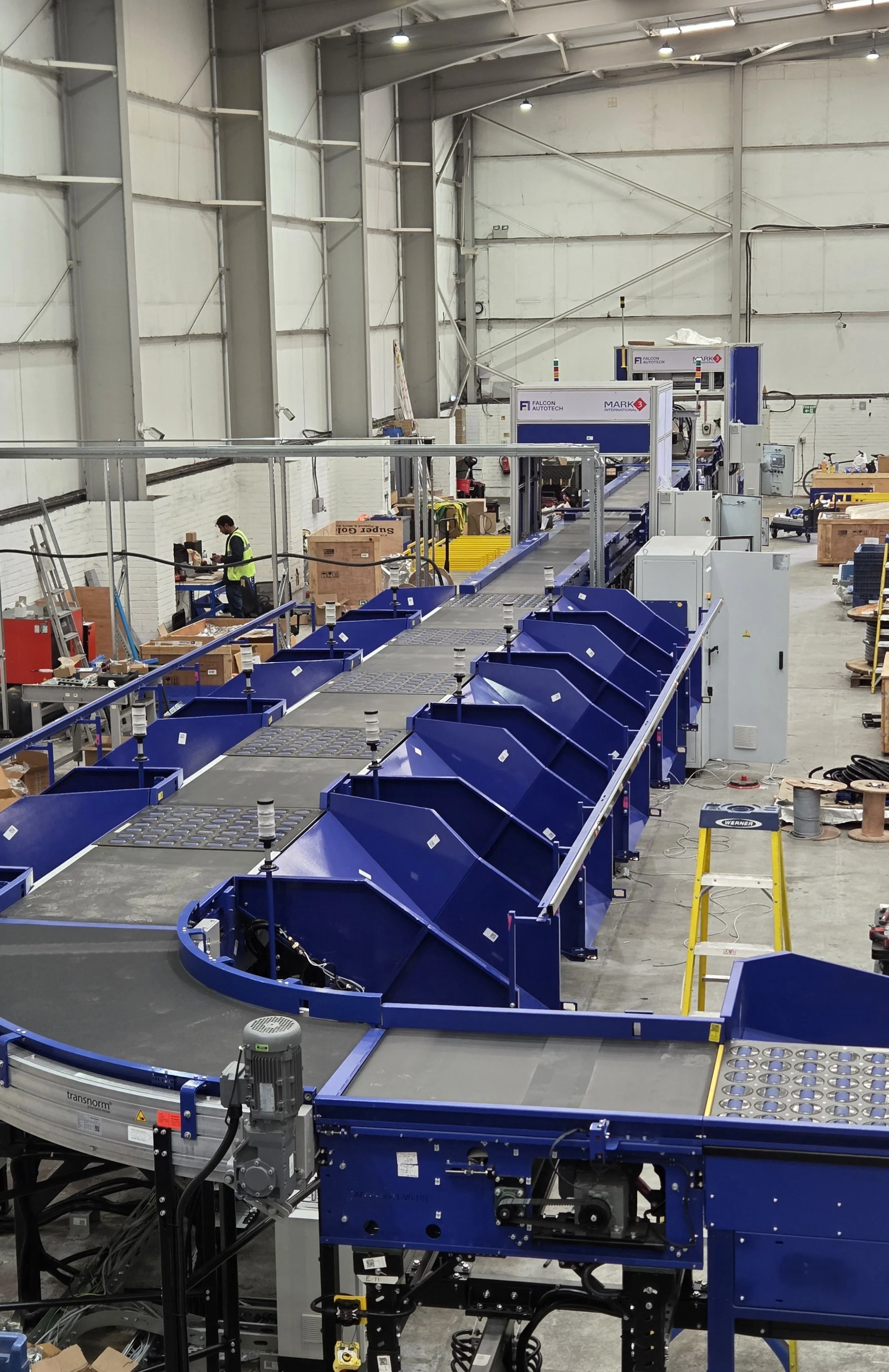 Industrial warehouse with the Mark 3 International blue conveyor system and machinery, worker in safety vest and helmet working among equipment and boxes.