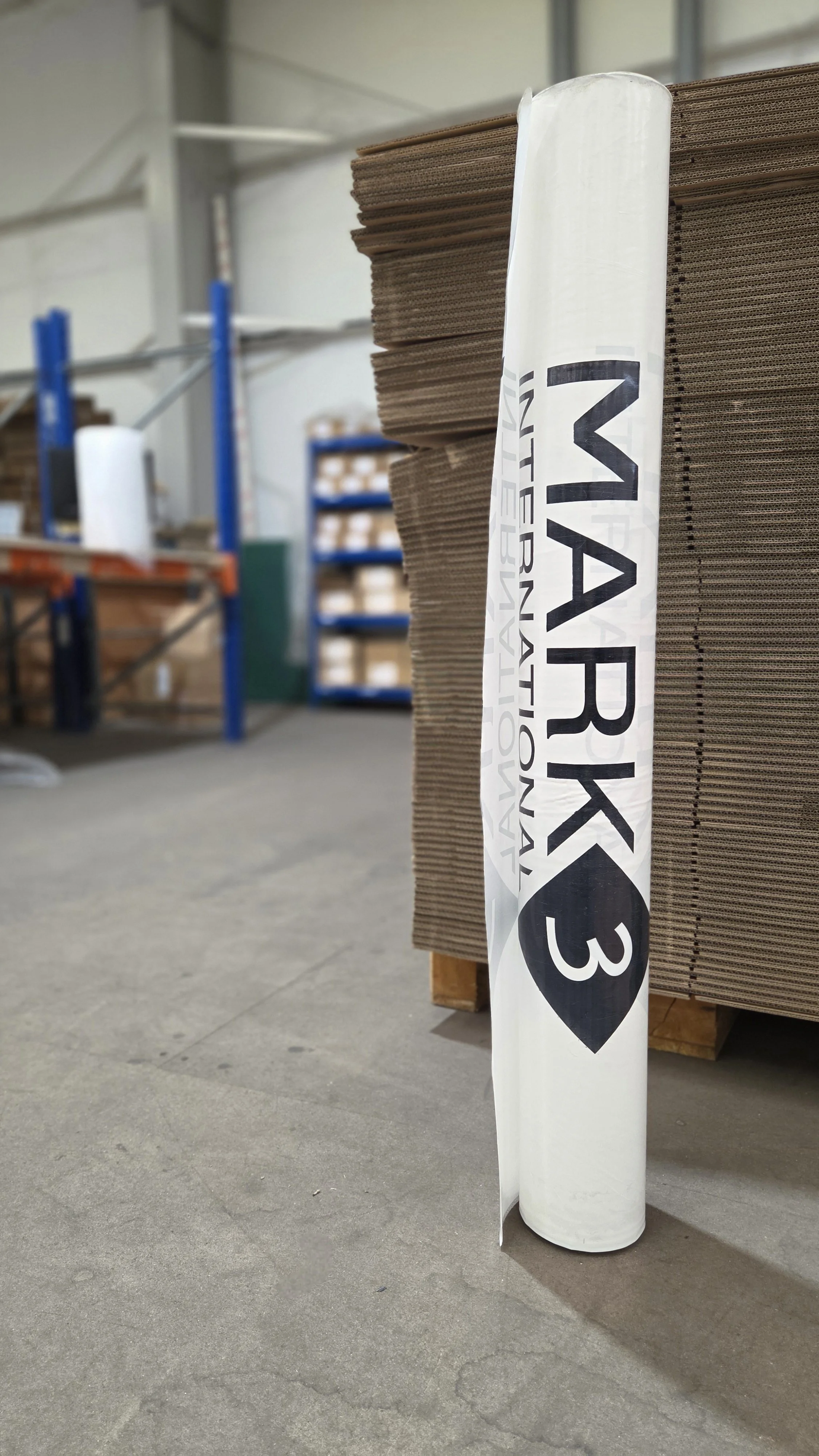 Large roll of plastic sheeting with the words 'Mark 3 International' partially visible, set in an industrial warehouse with stacked cardboard sheets and metal shelving in the background.
